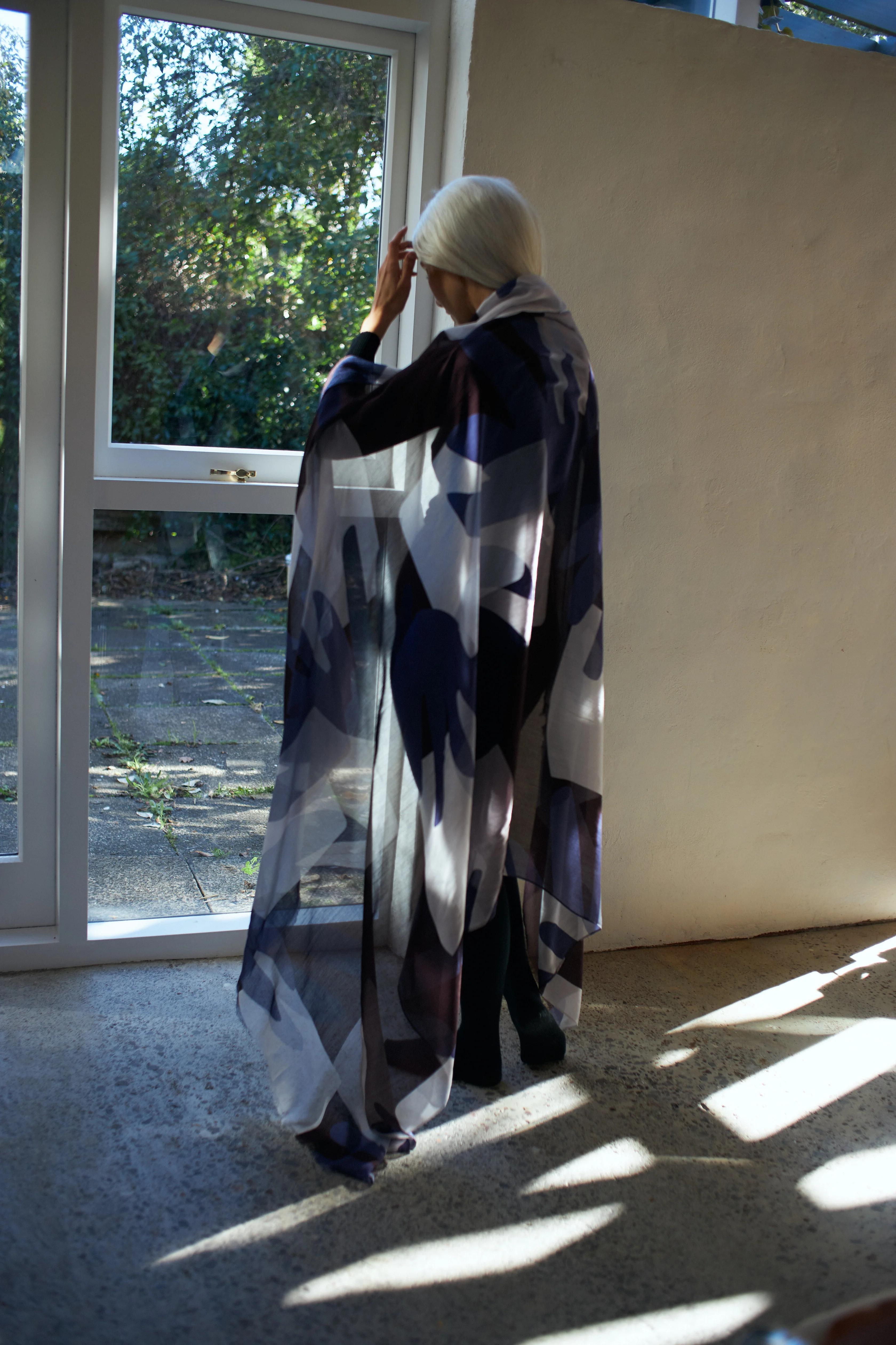 A person with long white hair stands indoors near a sunlit window, adjusting their hair. They are wearing a flowing, semi-transparent cape from Walker & Bing, adorned with abstract, geometric patterns in shades of navy and white. The outside view shows greenery and a patio that complements this textile design masterpiece.