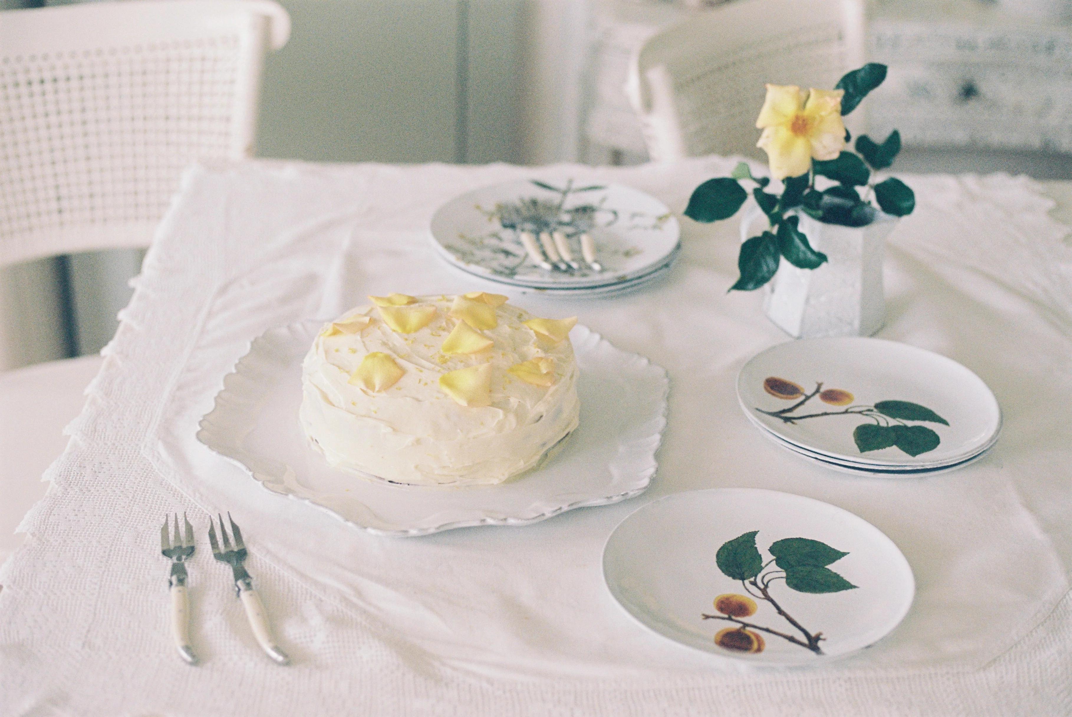 Frosted carrot cake with yellow flowers on scalloped platter. Dessert plates, forks, yellow rose vase nearby.