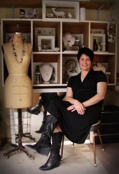 Jewellery designer in black outfit sits by vintage dress form with necklace. Shelves display organic-inspired collections, photos, and decor items.