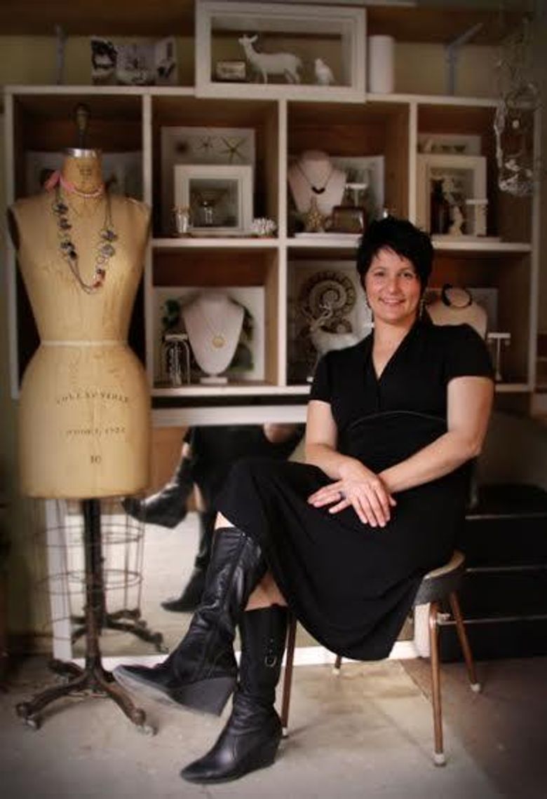 Jewellery designer in black outfit sits by vintage dress form with necklace. Shelves display organic-inspired collections, photos, and decor items.