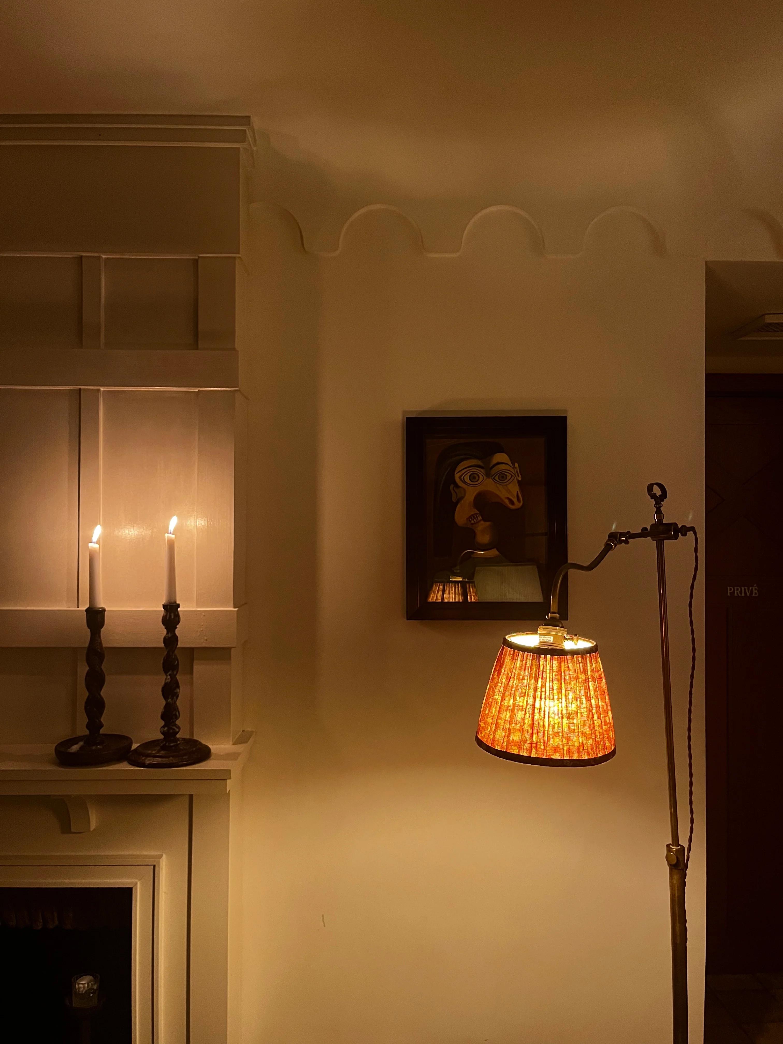 Dimly lit room with lit candles on shelf, framed Paris painting, and orange lampshade floor lamp.