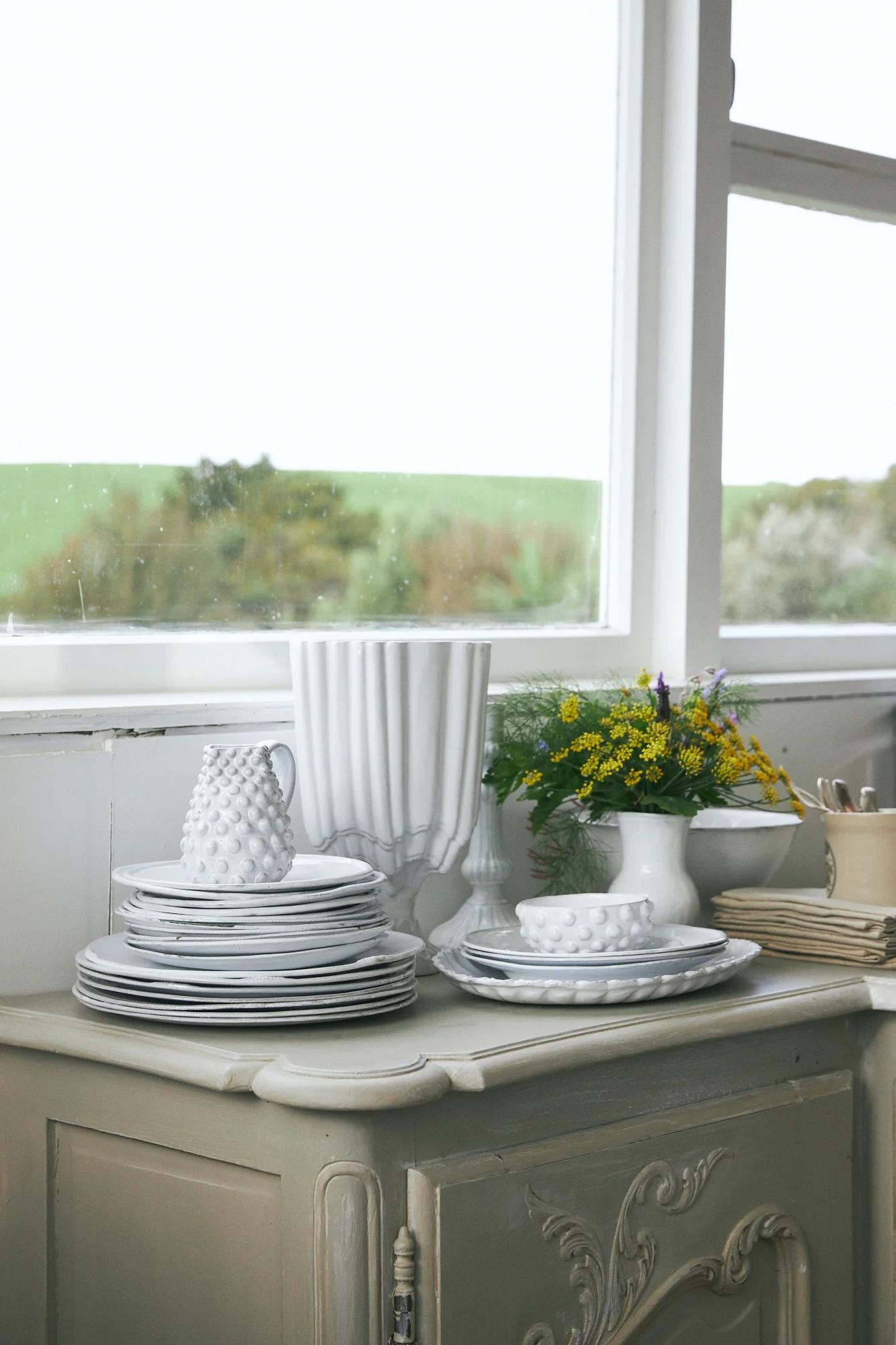 White plates, decorative pitcher, and vase with flowers on vintage cabinet. Window shows green fields view.