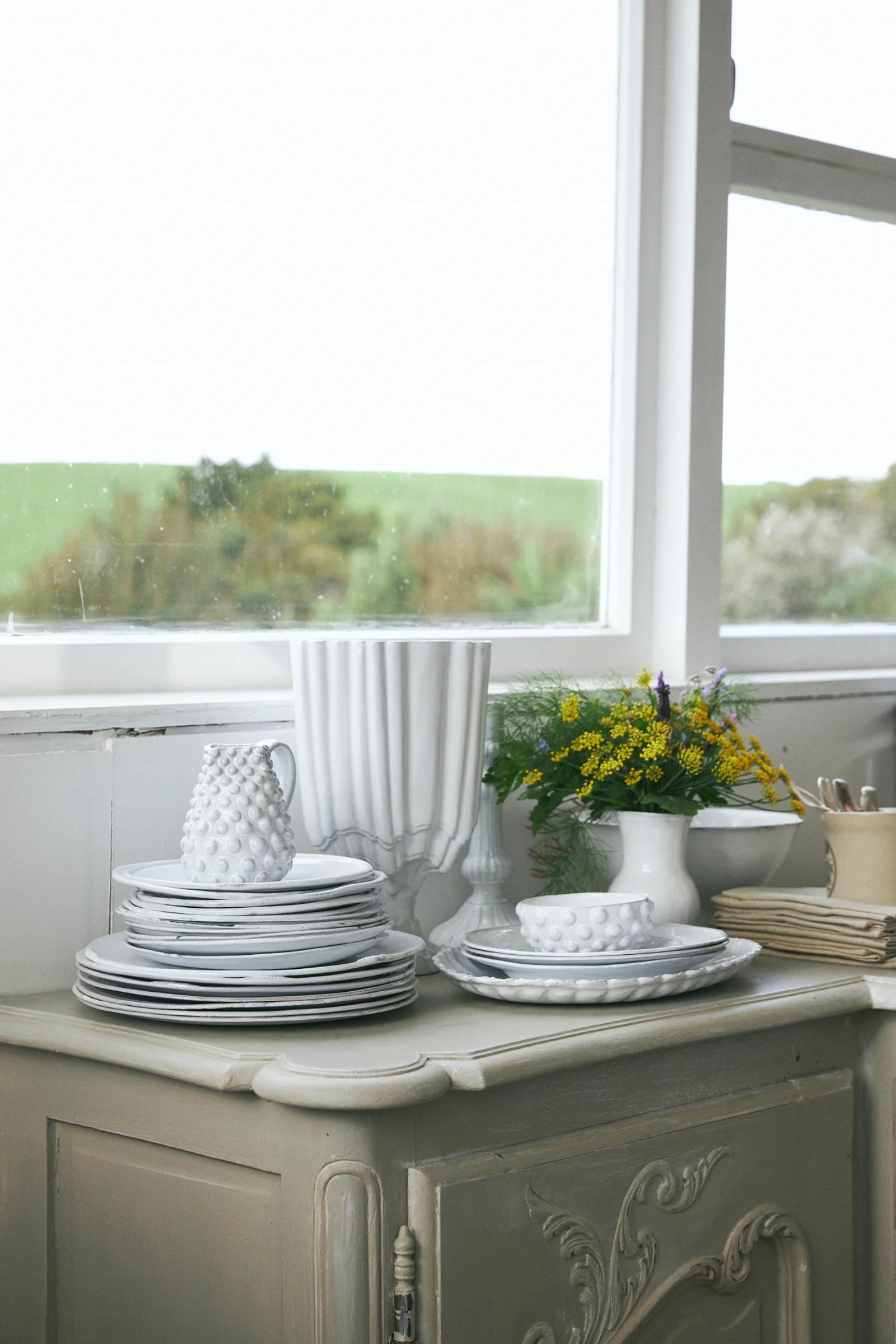 White plates, decorative pitcher, and vase with flowers on vintage cabinet. Window shows green fields view.