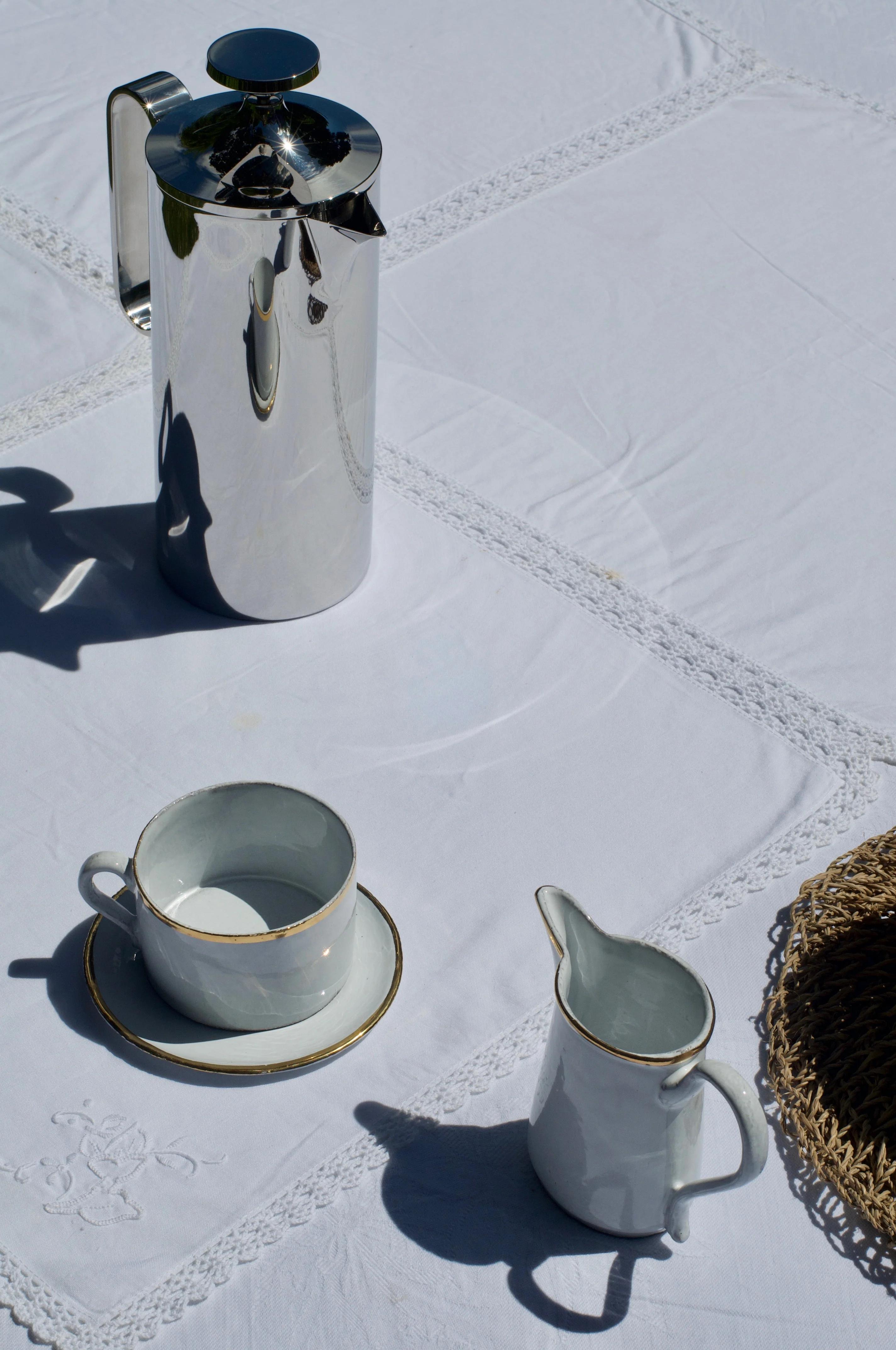 Steel coffee pot, white gold-trimmed teacup, saucer, and creamer on lace-edged tablecloth. Partial view of woven placemat.