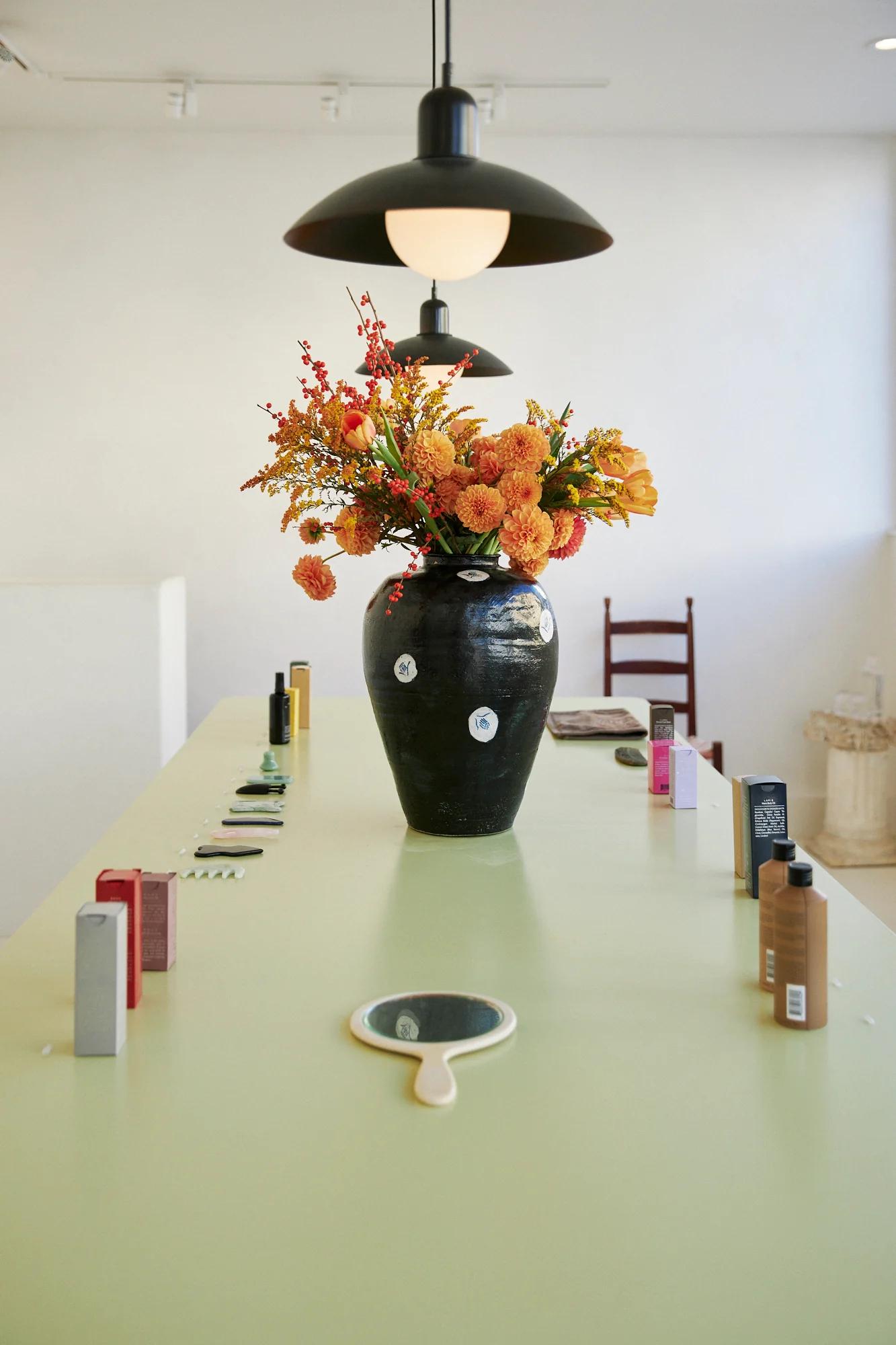 Green table with skincare products and perfumes. Vase with orange and yellow flowers. Small mirror in front. Chair in background.