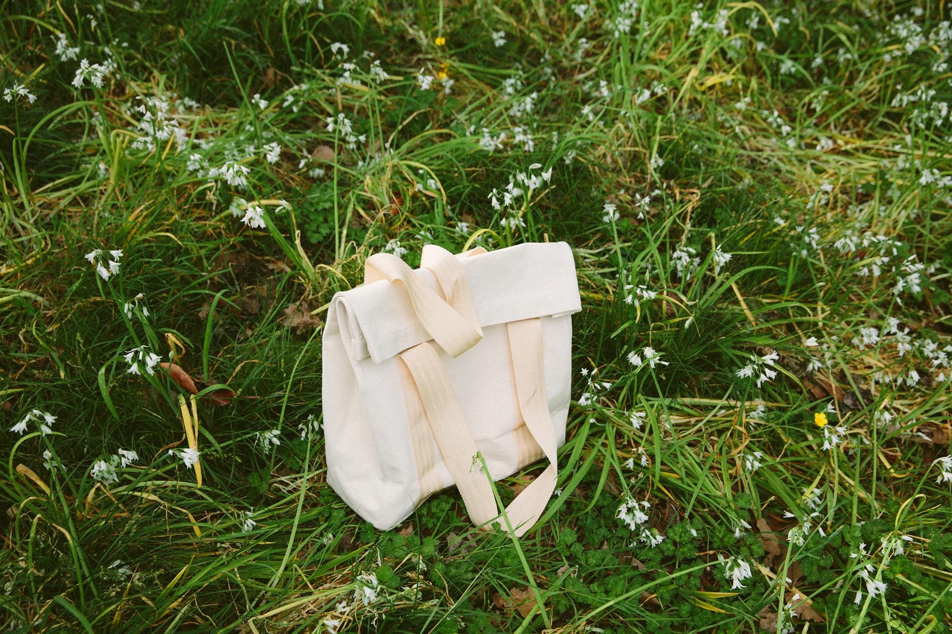 Beige canvas backpack with cream straps on grass amid small white flowers. Simple, folded-top design by KLAY blends with natural setting.