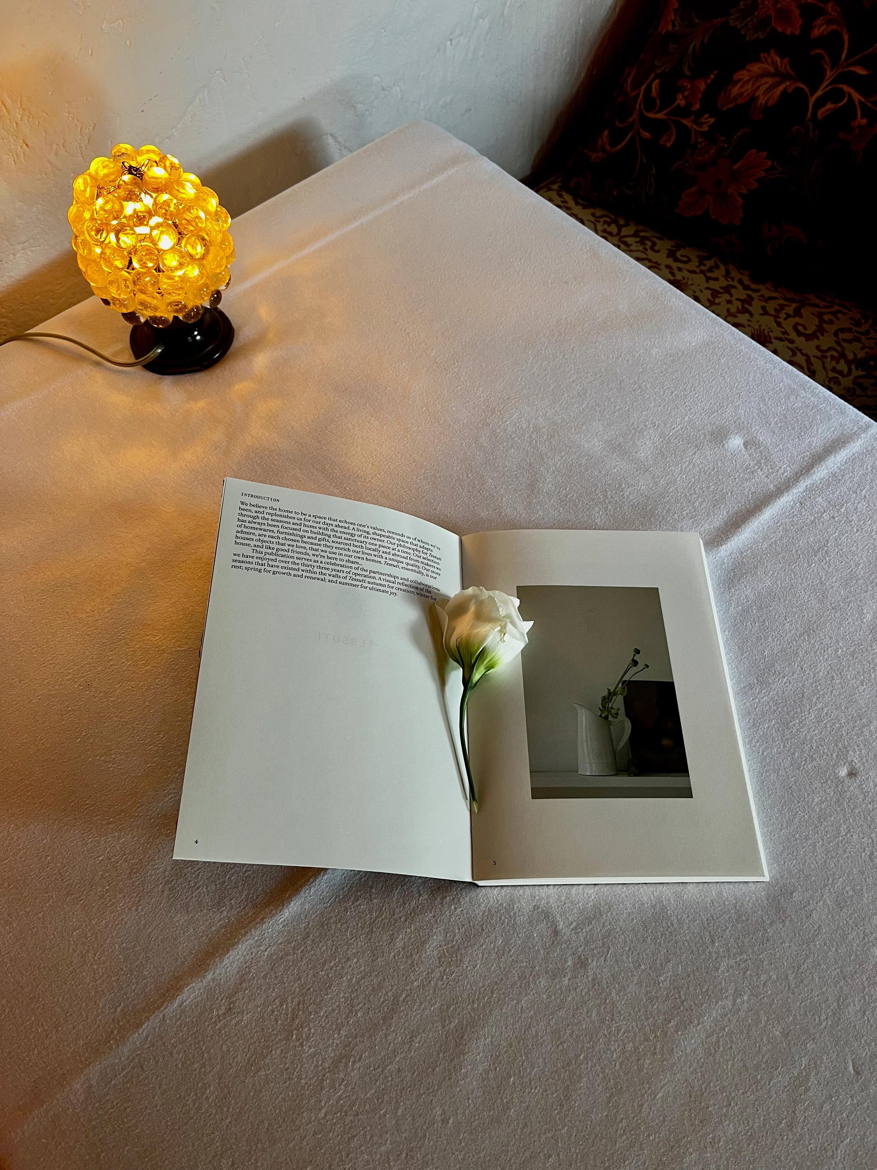 Small lamp with yellow shade on white-clothed table. Open book with rose on pages.