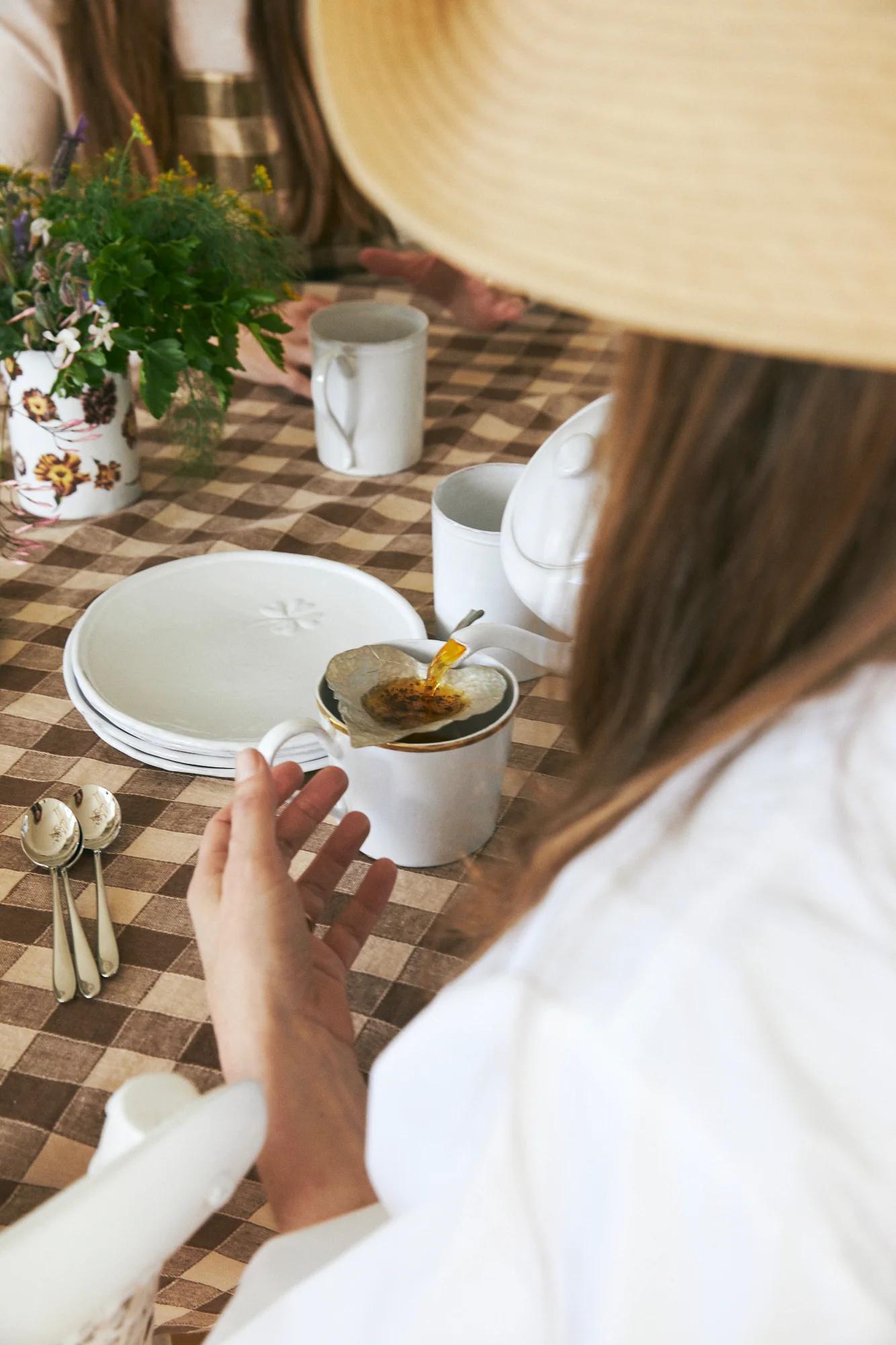 Person in wide-brimmed hat at checkered table, holding tea. Table set with plates, cups, flowers. Another person seated across.