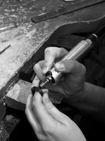 Black and white image: Jewellery designer uses power tool on small object in cluttered workspace.