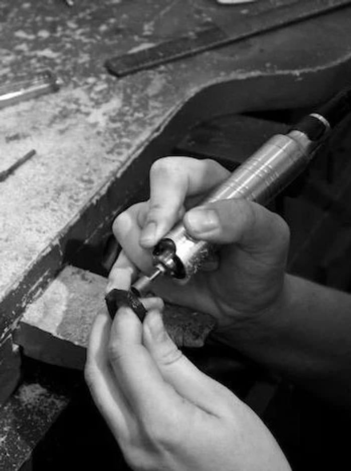 Black and white image: Jewellery designer uses power tool on small object in cluttered workspace.