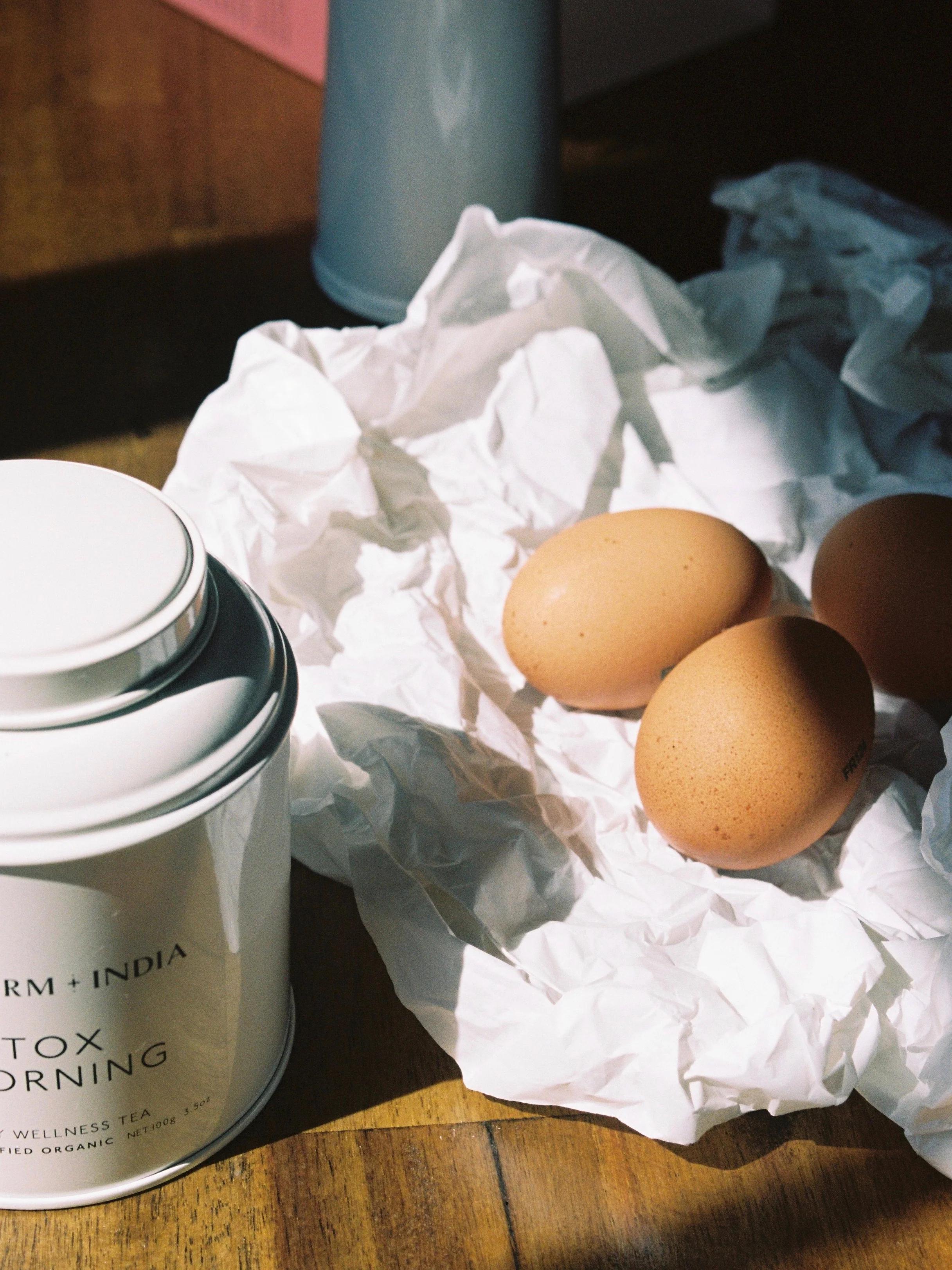 A white canister beside brown eggs on crumpled paper, lit by natural sunlight on a wooden table, creating a warm baking scene.