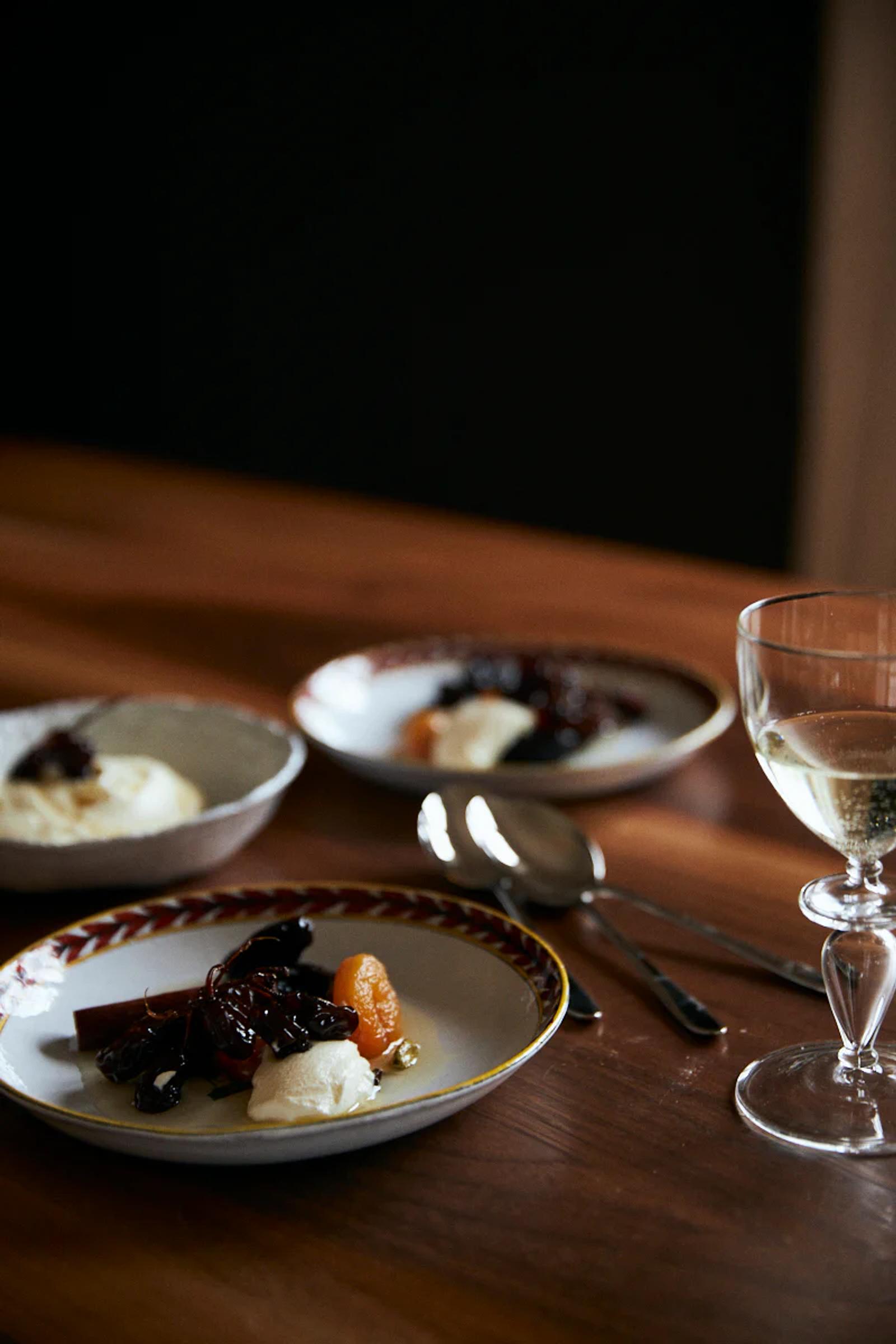 Three plates with cream and dark vegetable pieces on wooden table. Wine glass, fork, and spoon nearby. Dimly lit background.