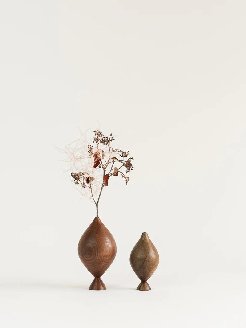 Two wooden vases of different sizes are set against a neutral background. The larger vase holds dried branches with small flowers, while the smaller vase remains empty. Both have a smooth, tapered finish.