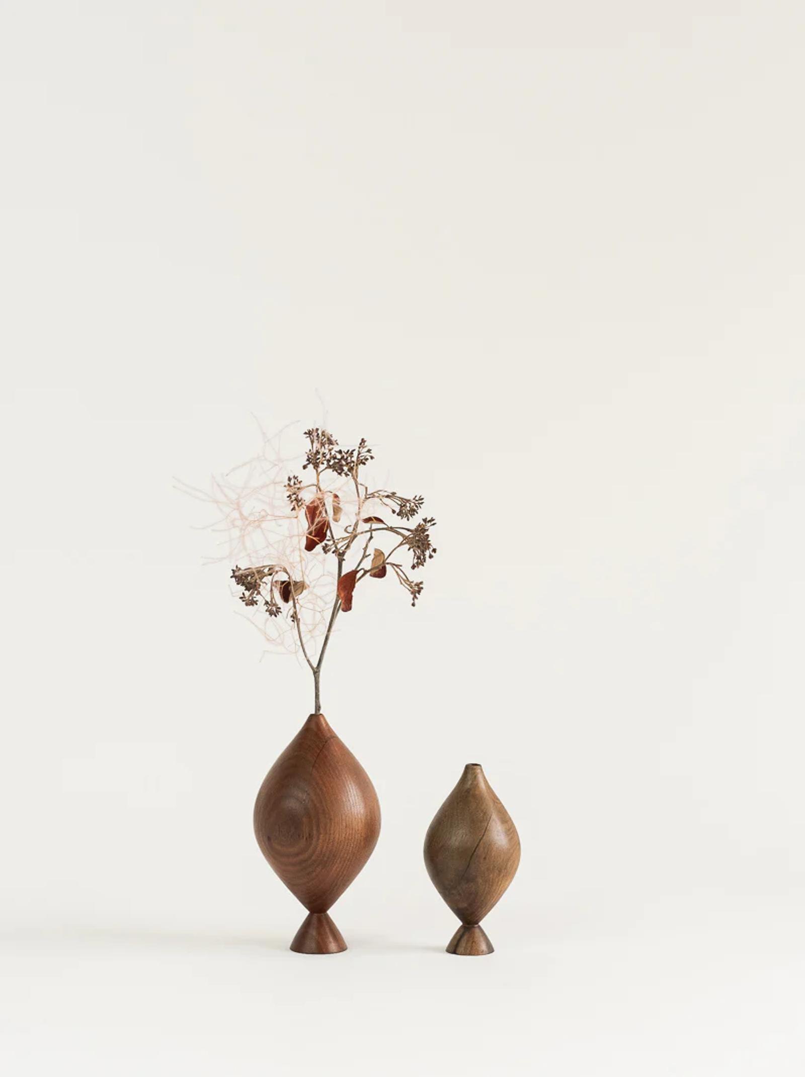 Two wooden vases of different sizes are set against a neutral background. The larger vase holds dried branches with small flowers, while the smaller vase remains empty. Both have a smooth, tapered finish.