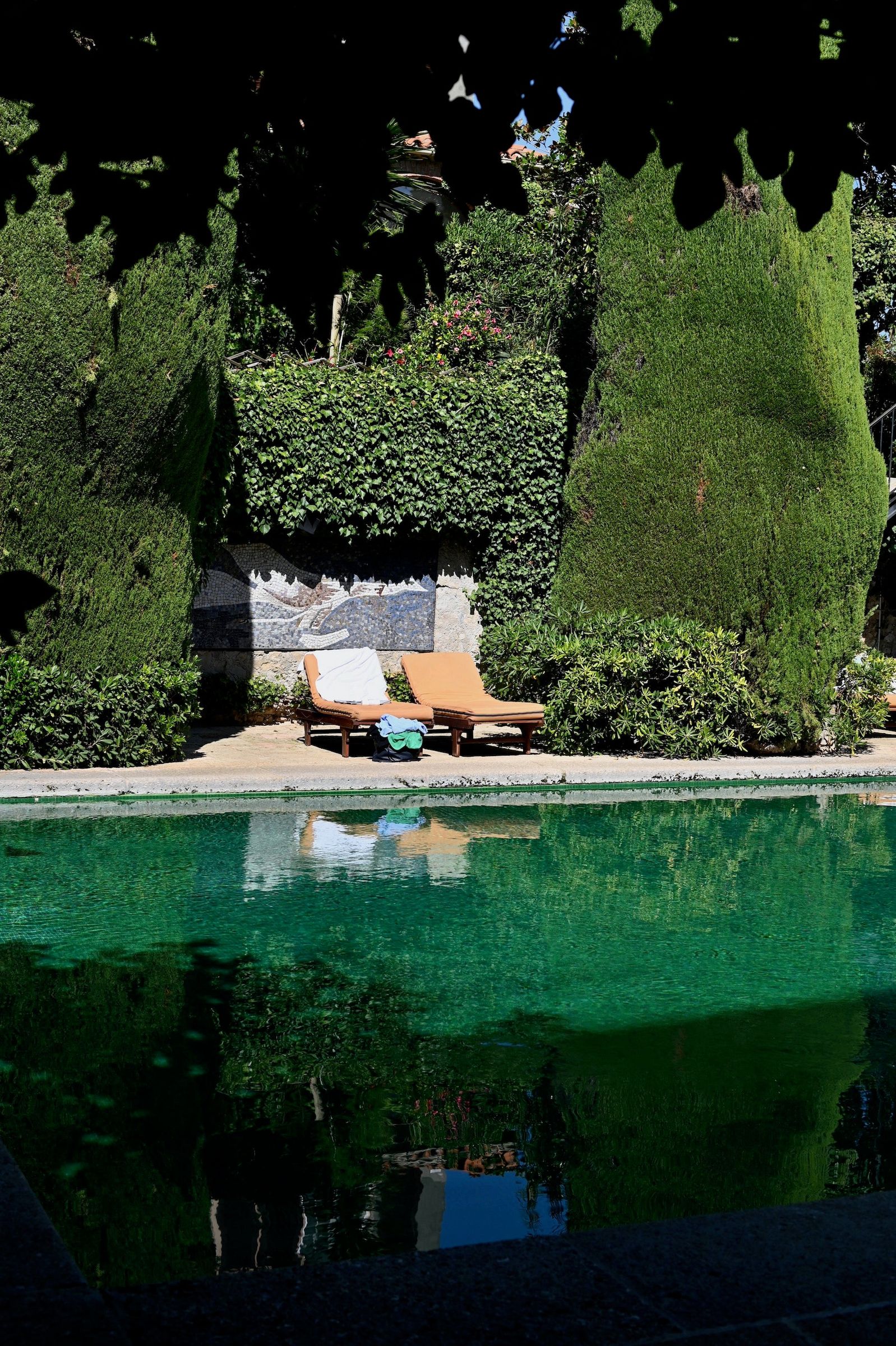 Two empty lounge chairs by green pool, surrounded by tall hedges. Secluded atmosphere with lush foliage casting shadows.