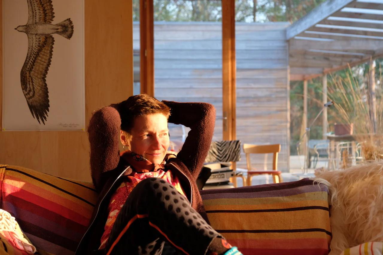 Smiling person on striped sofa in sunlit room with wooden walls. Bird illustration on wall, large windows with outdoor view.