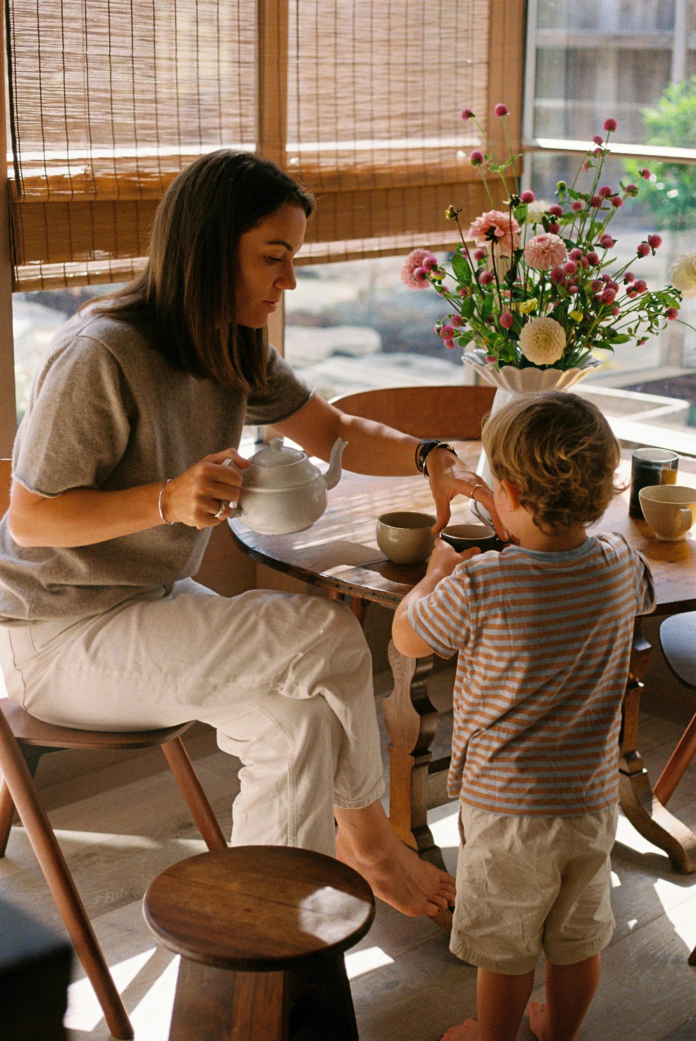 Woman pours tea for young boy in sunlit room. Wooden table with flower vase. Boy in striped shirt, woman in casual attire.