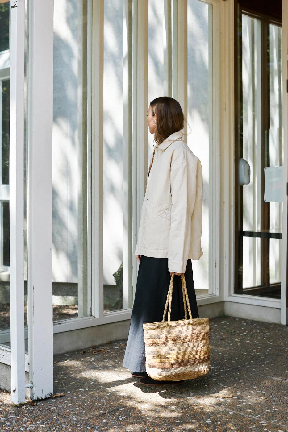 Person with long hair outside glass building. Holding striped bag, looking away. Tree shadows on glass.