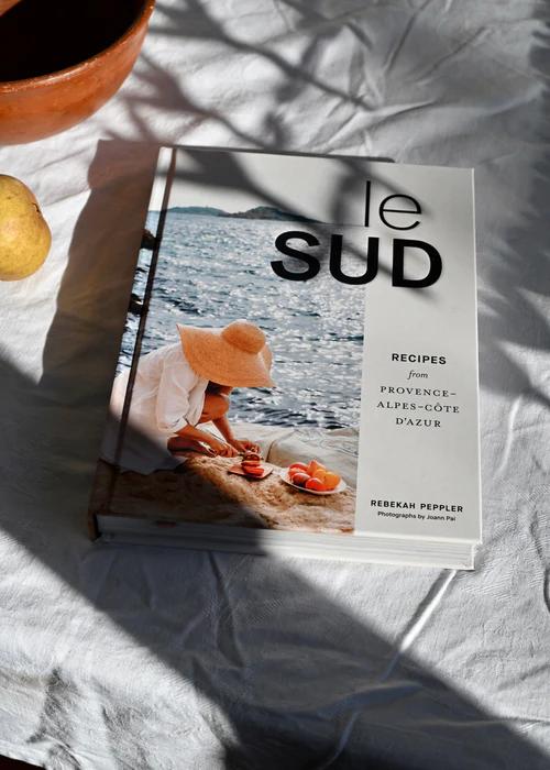 "Le Sud" cookbook on beige surface. Cover shows person in hat by sea arranging produce on beach.