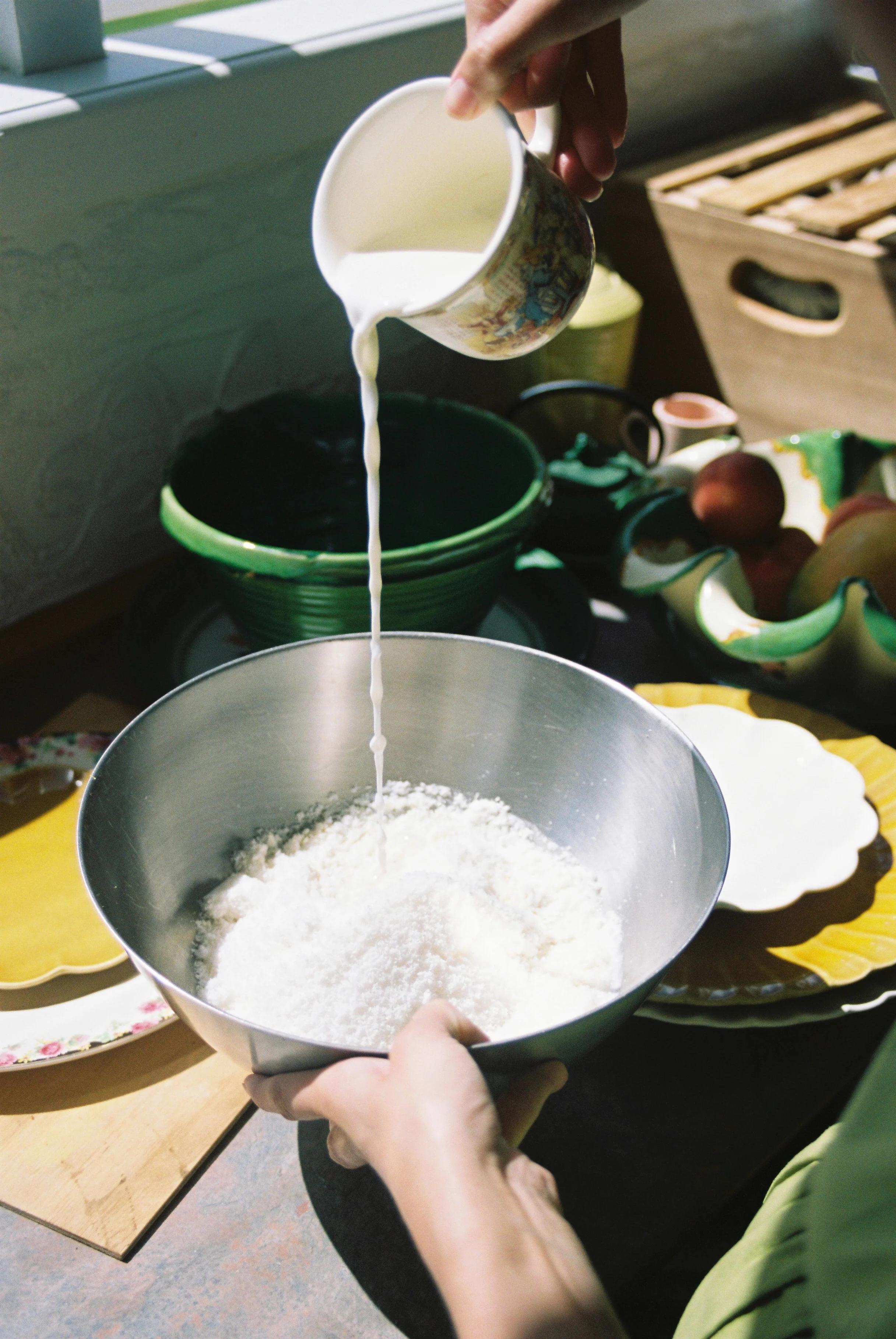 Person pouring milk from floral jug into mixing bowl with flour. Sunlit kitchen counter with dishes and fruit.