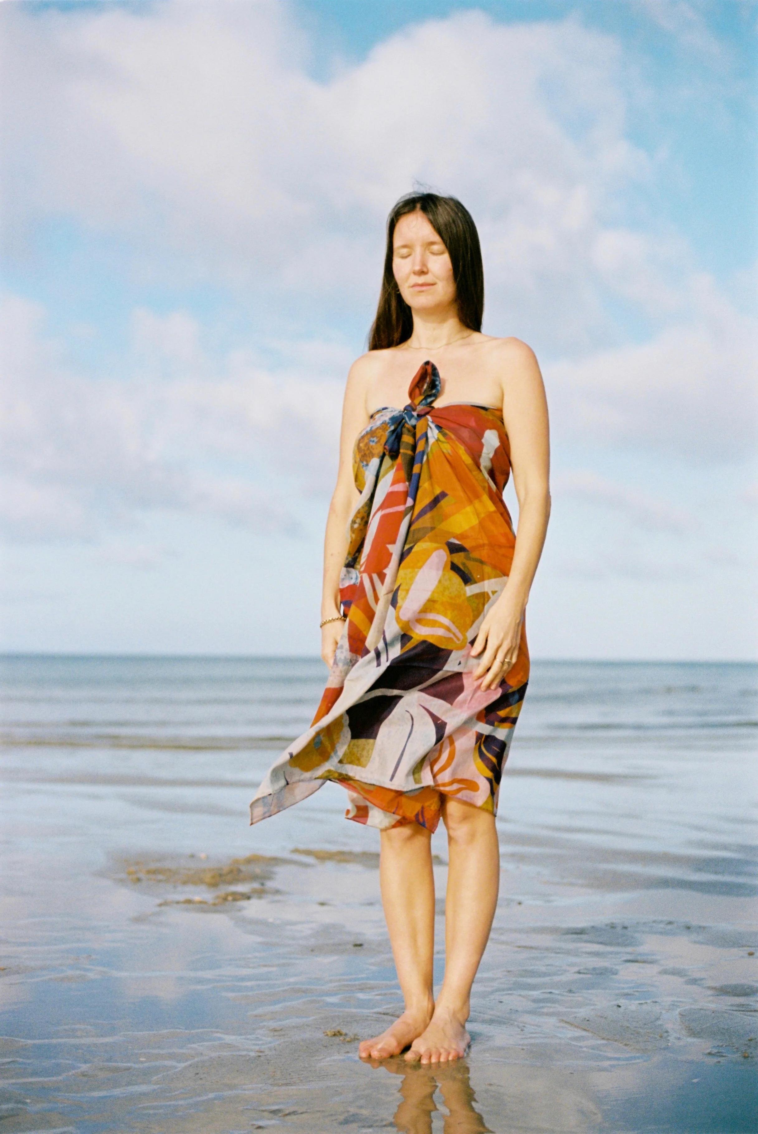 Woman in colourful patterned wrap dress on beach with eyes closed. Long hair, partly cloudy sky, calm ocean in background.