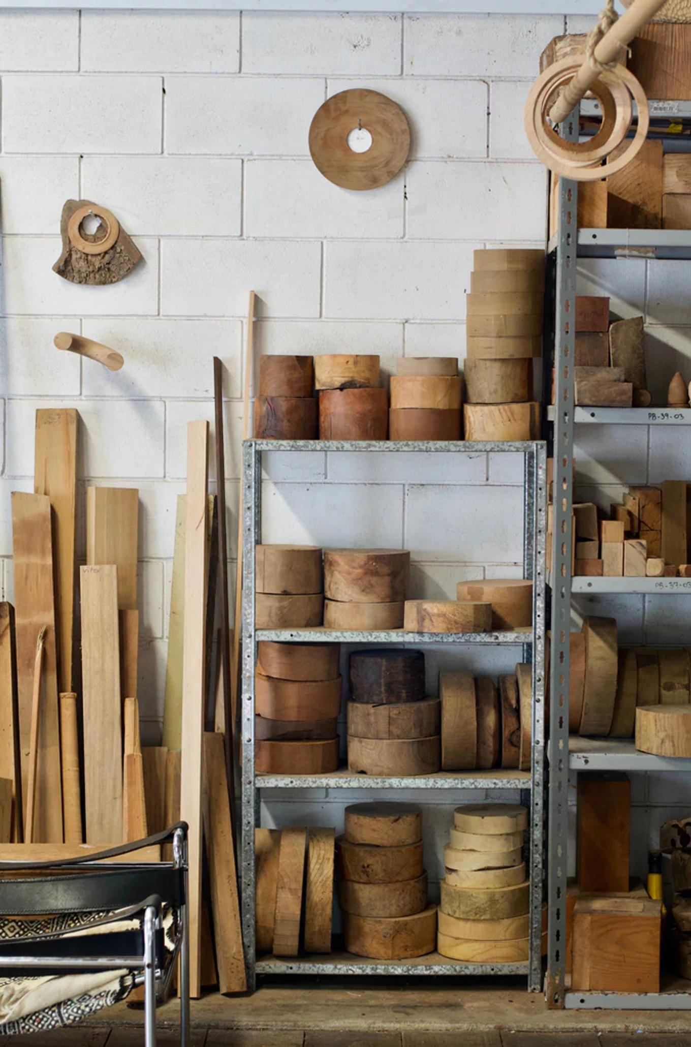 Workshop with tall shelves holding wooden logs and materials. Various wood planks lean against brick wall, chair nearby.