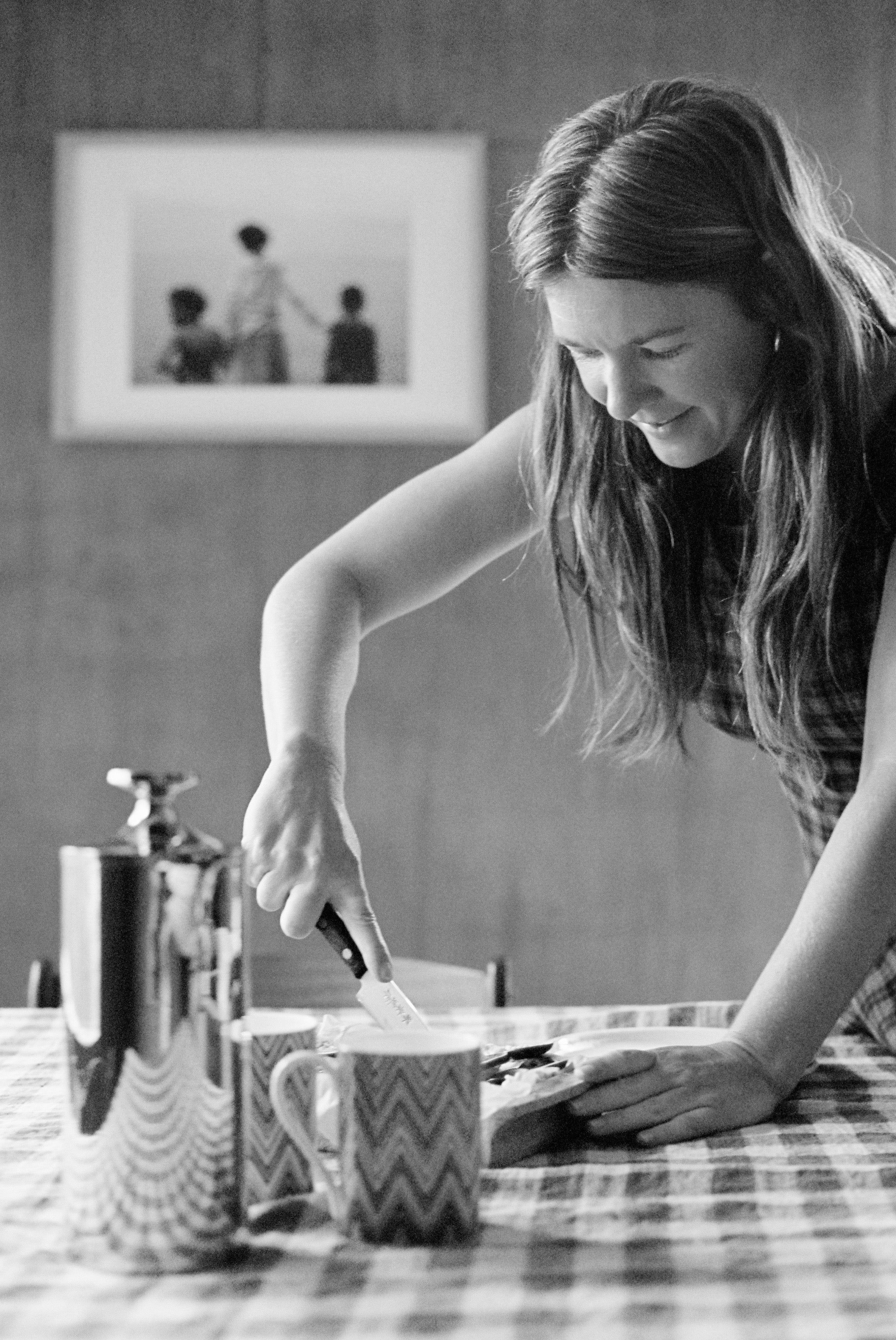 Smiling woman spreading food on bread. Checkered tablecloth, framed photo behind.