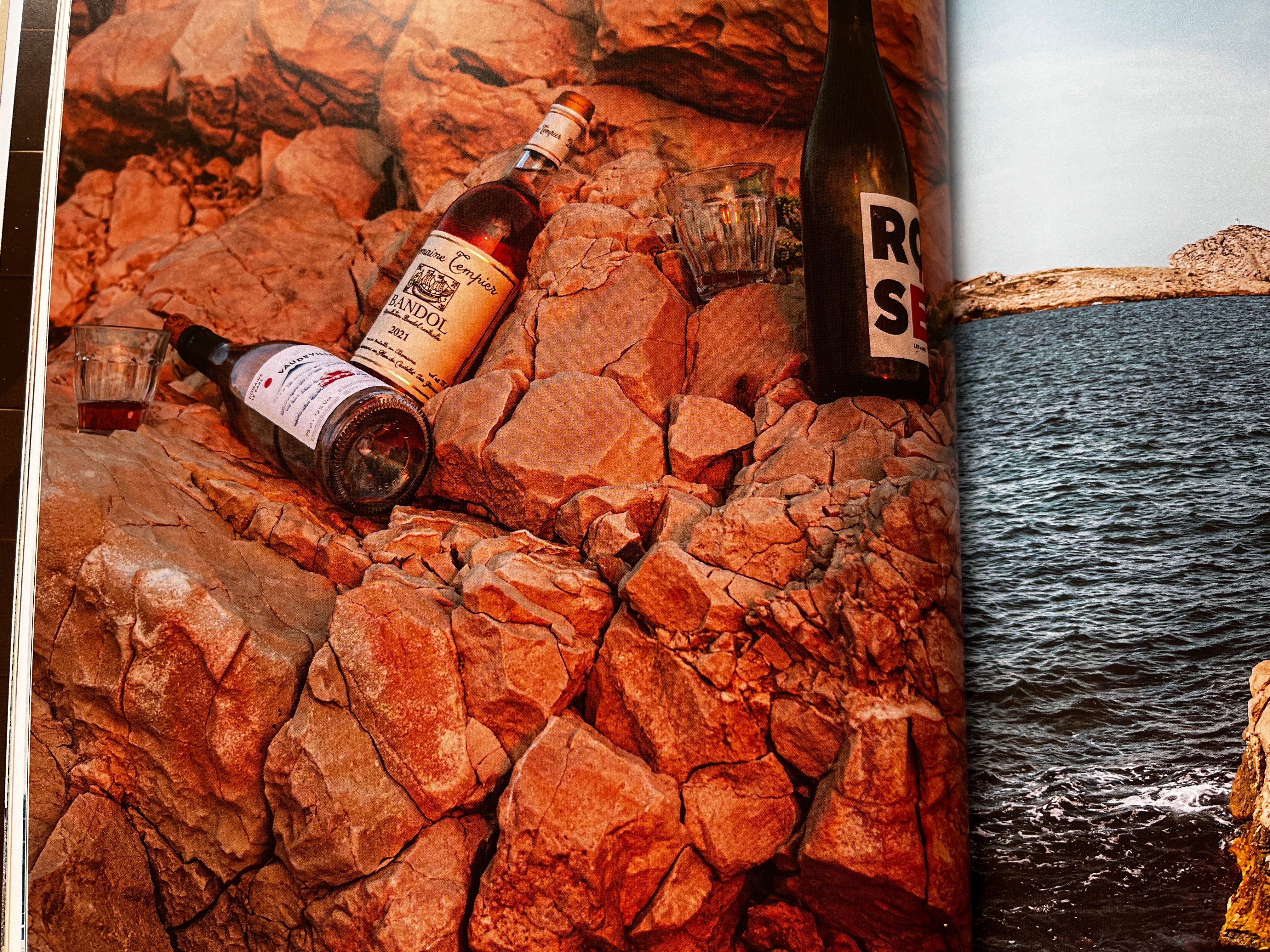 Three bottles and glasses on rocks near water. Warm, reddish hue. Rocky hills and water in background.