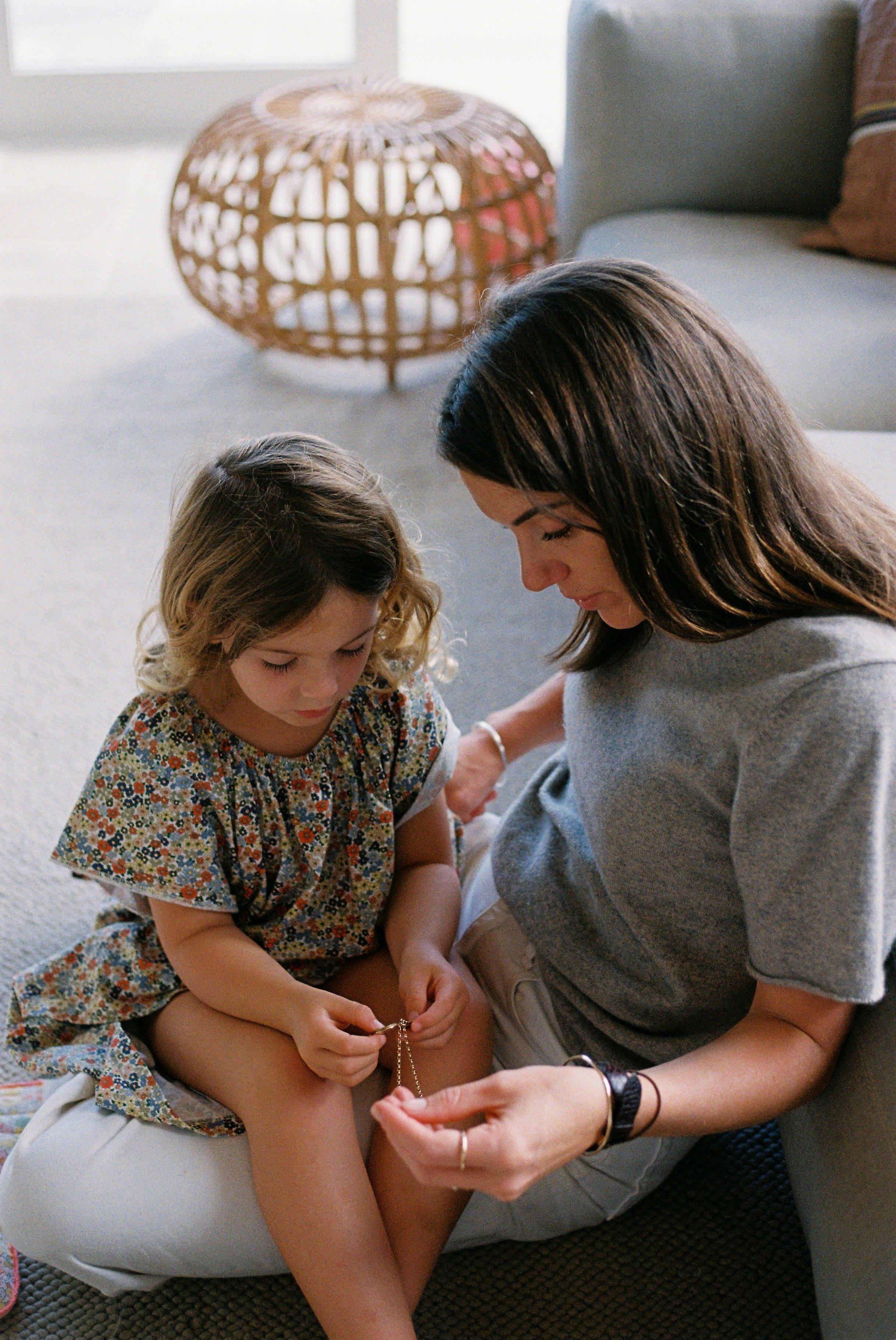 Woman on sofa watches young girl in floral dress examining small object. Wicker stool in background.