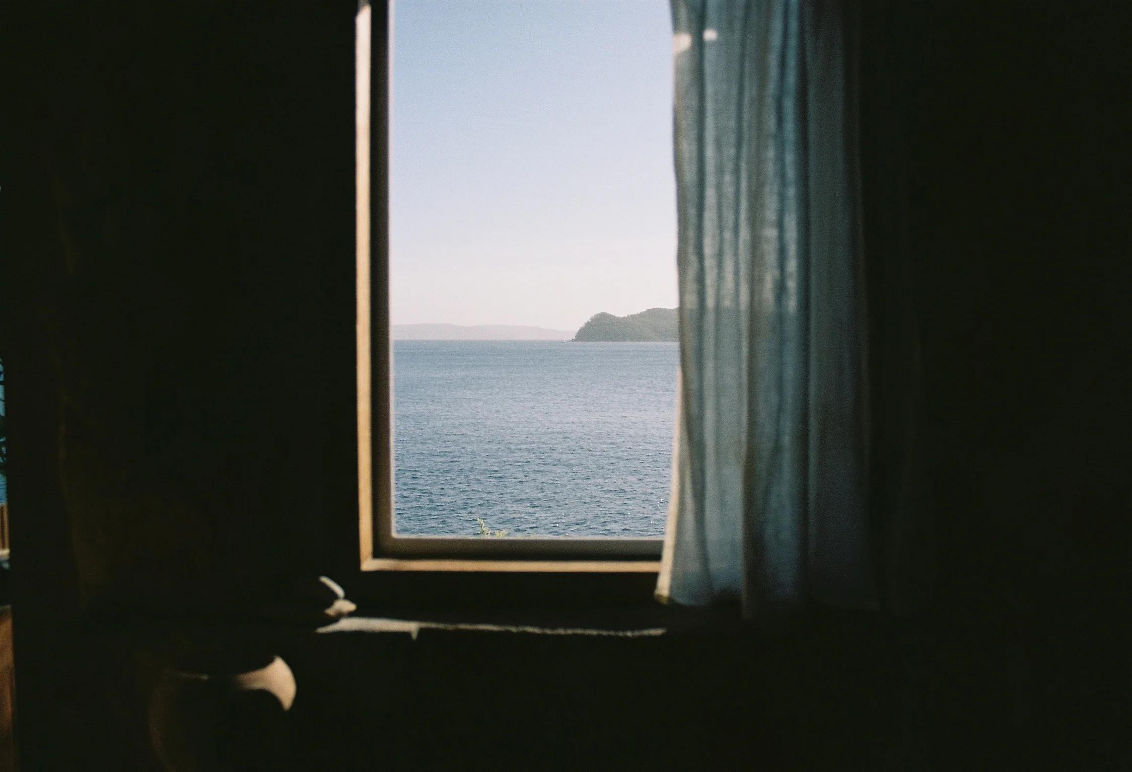 A serene sea view through a window framed by blue curtains. Calm waters stretch to a distant coast under a clear sky, capturing a peaceful moment.