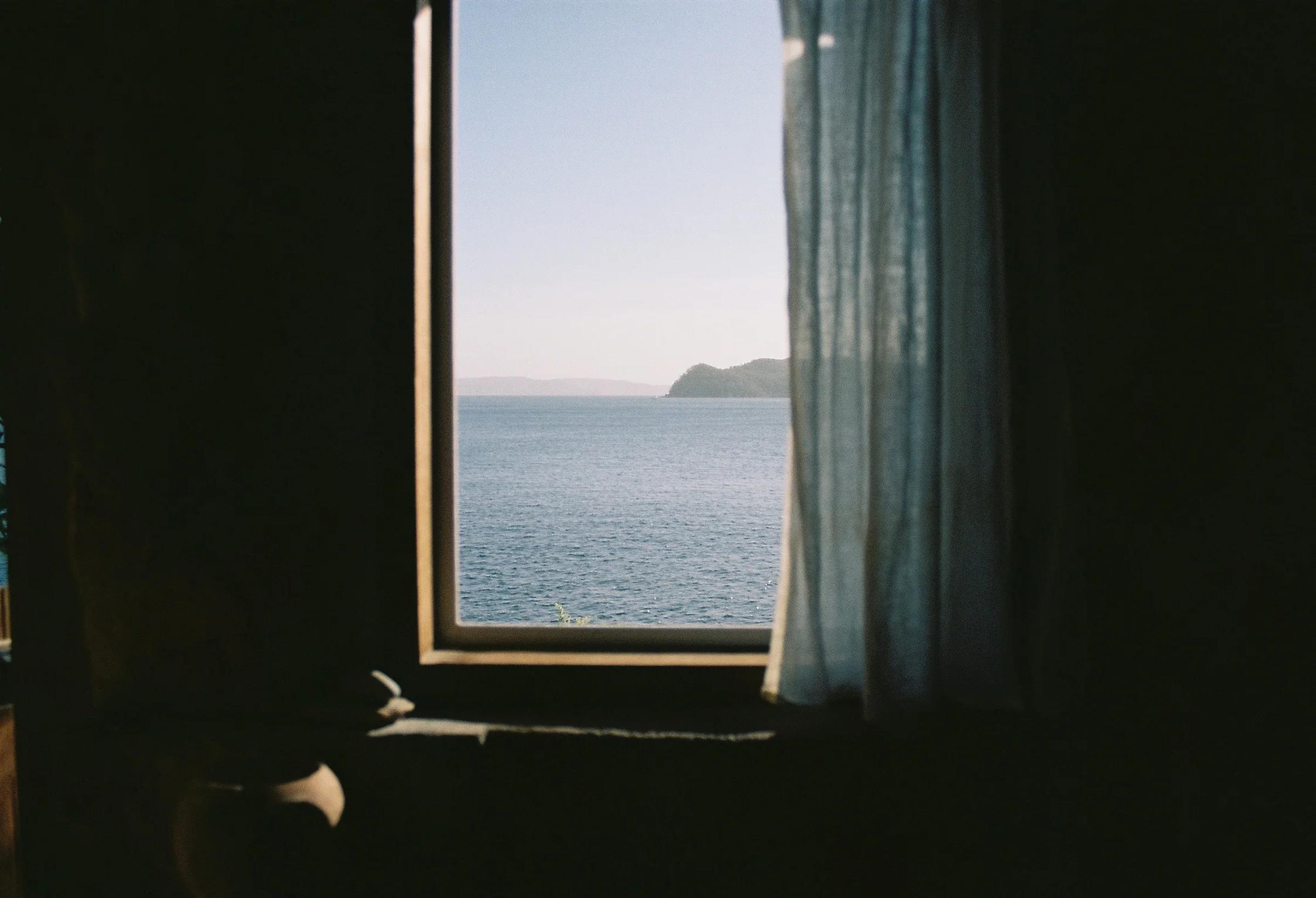 A view of calm ocean waters framed by a window in a seaside cottage, with light blue curtains partially drawn and a distant landform visible.