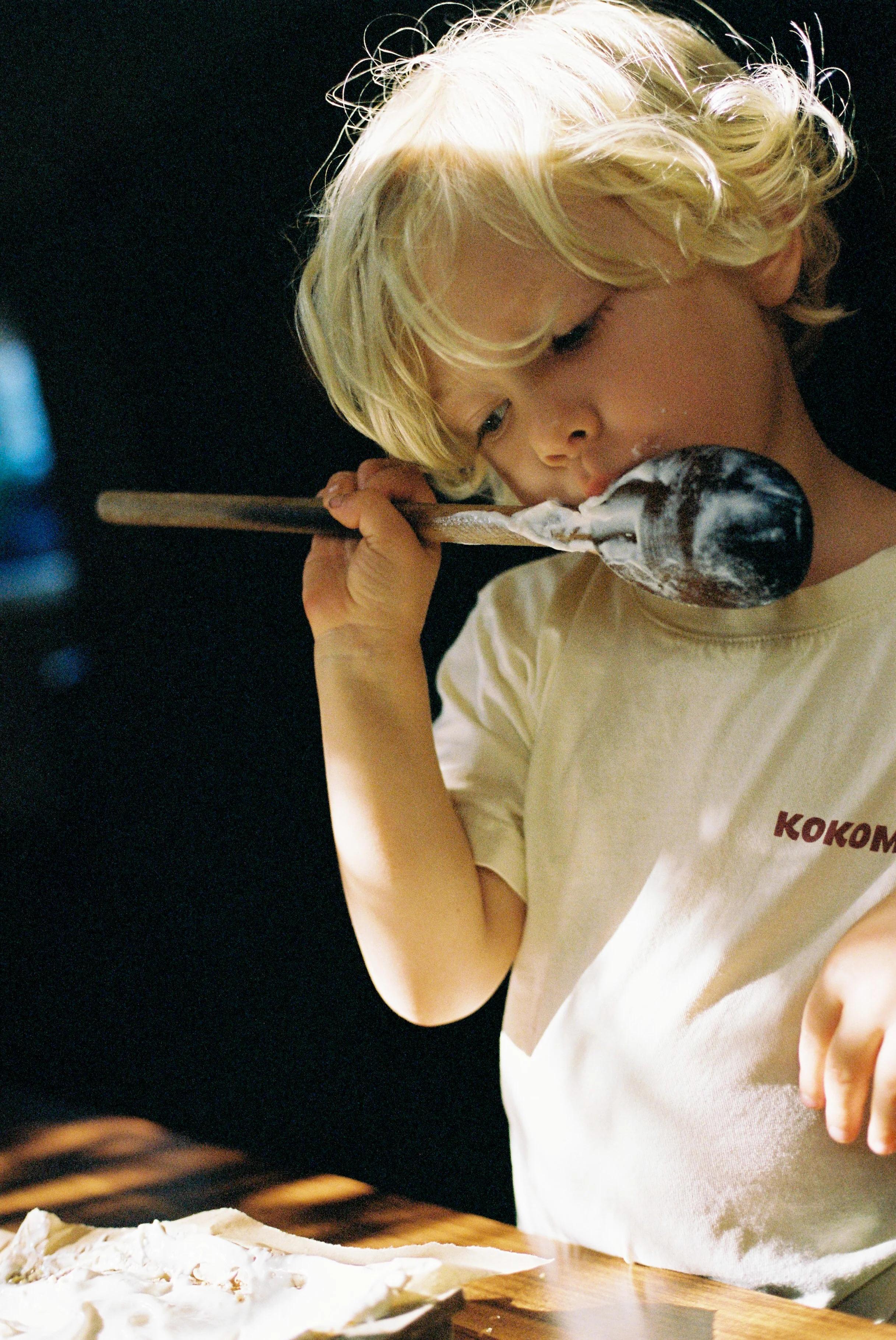 Child licking frosting-covered spoon. Soft lighting against dark backdrop.