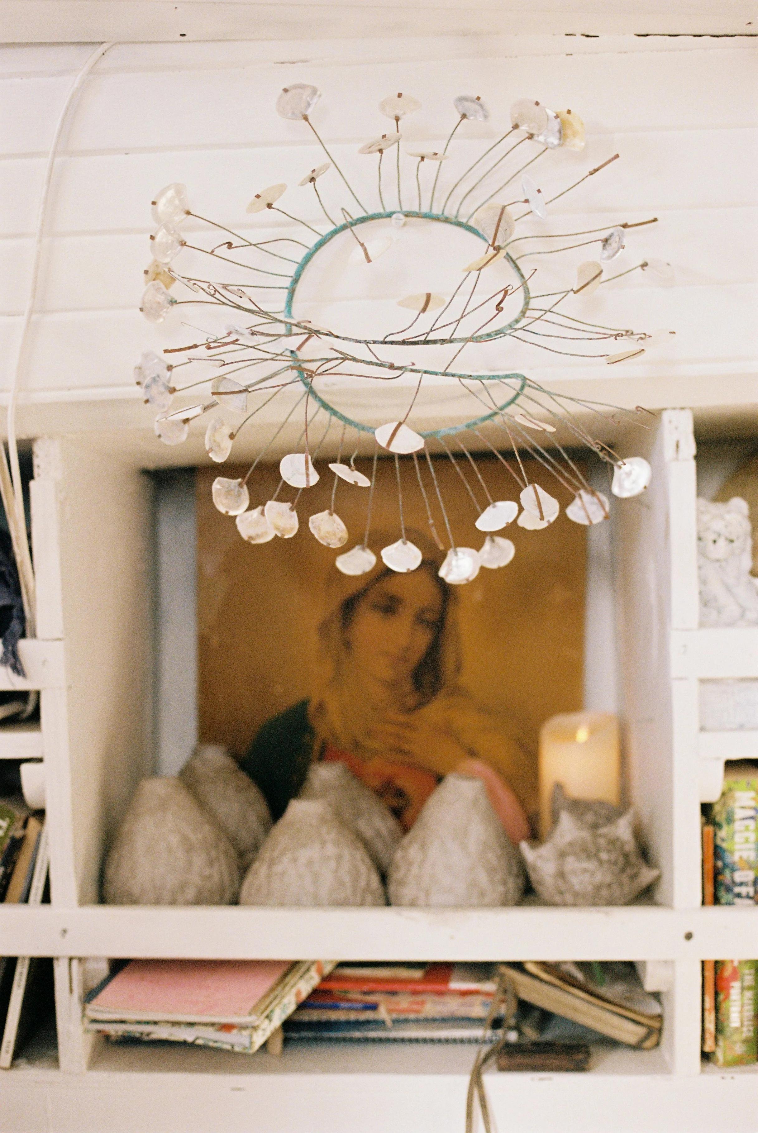 Shelf with framed painting, sculpted figures, lit candle, and books. Wire and shell mobile above.