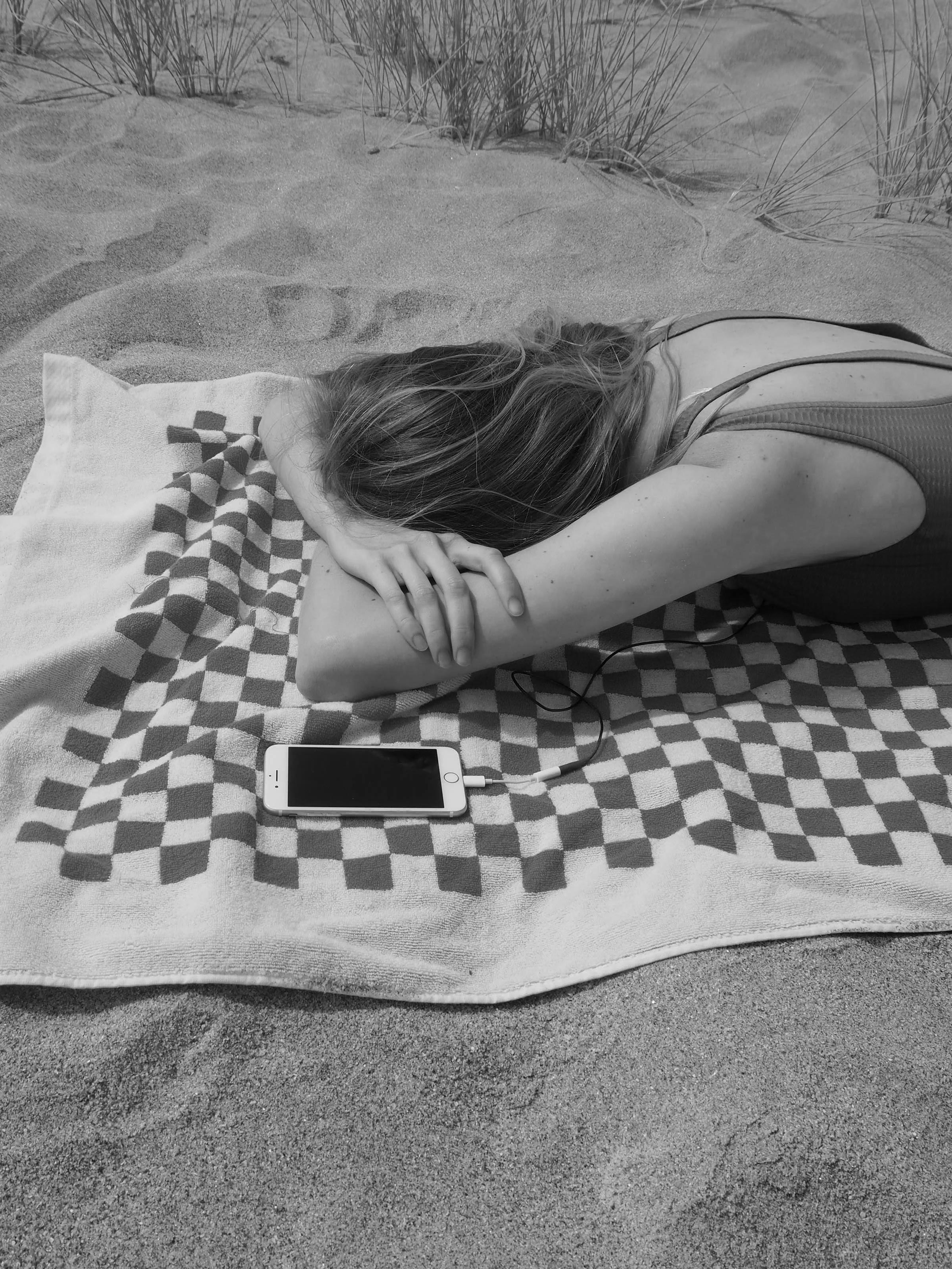 A woman lies face down on a checkered towel at the beach, with a smartphone beside her. The black-and-white background shows grains of sand and sparse grass.