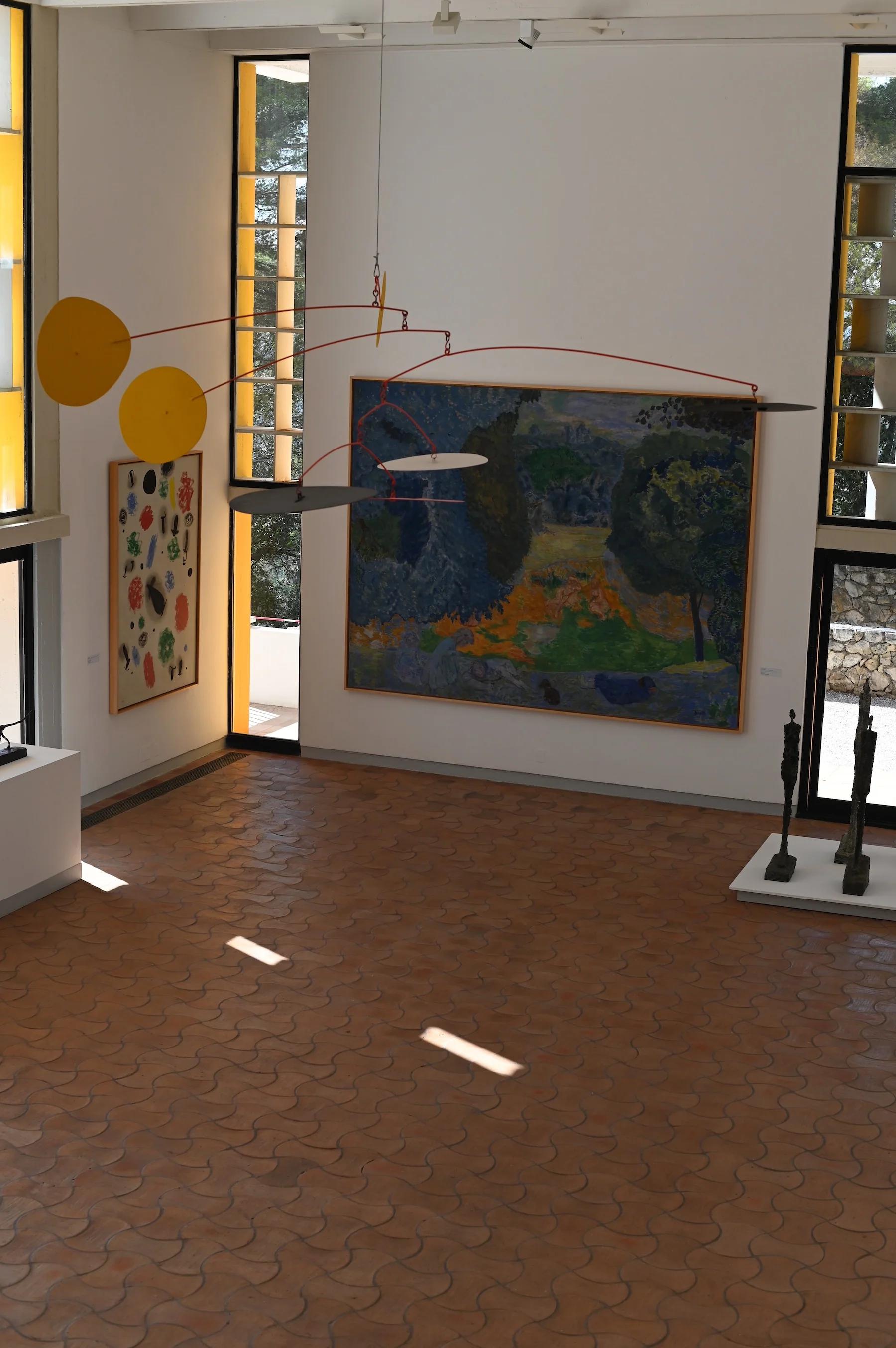 Art gallery with large landscape painting, abstract botanical work, and yellow mobile. Abstract sculptures near floor-to-ceiling windows.