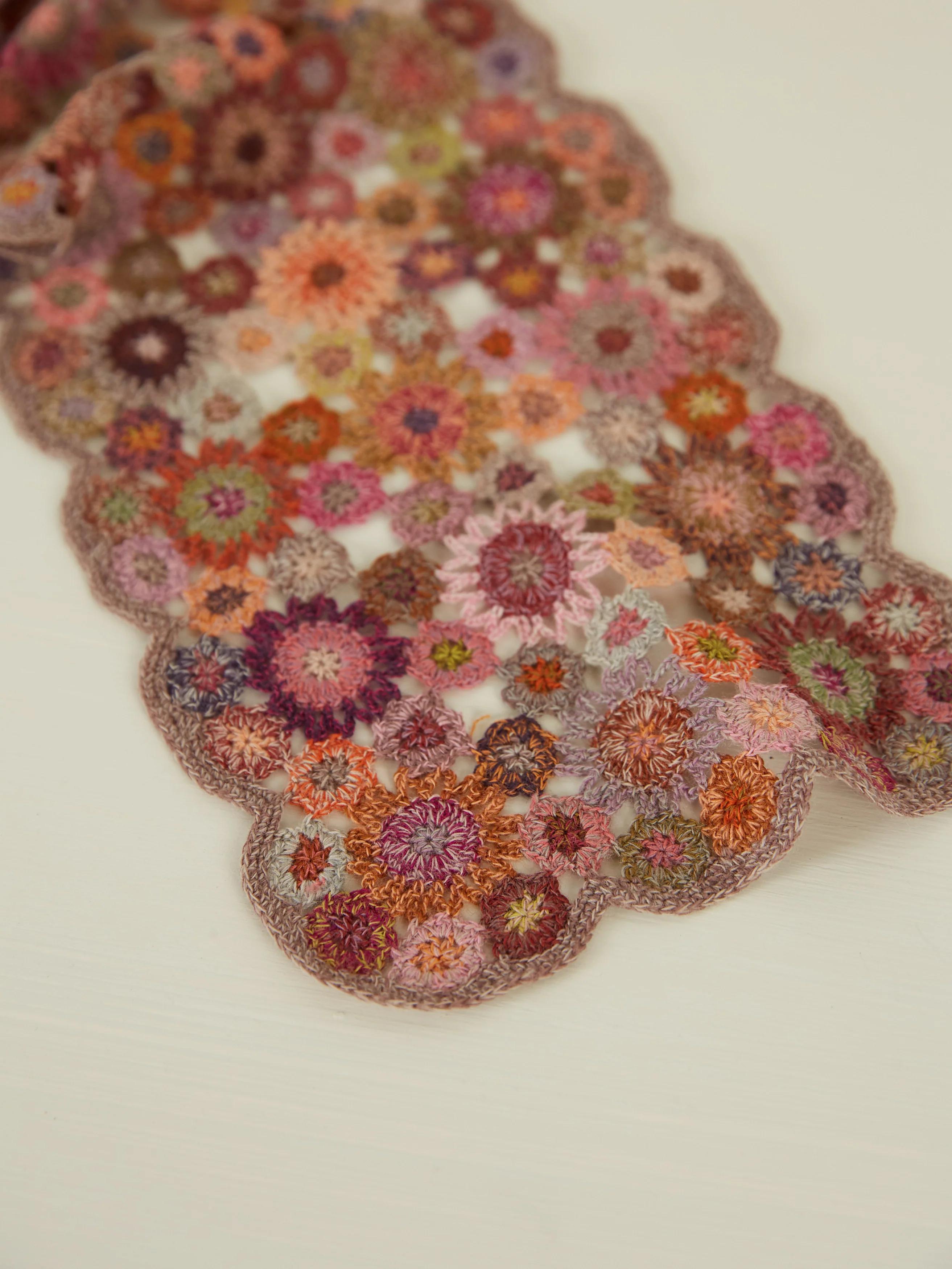 Close-up of crocheted flowers in pink, purple, orange, and brown, forming a scalloped edge against a white background.