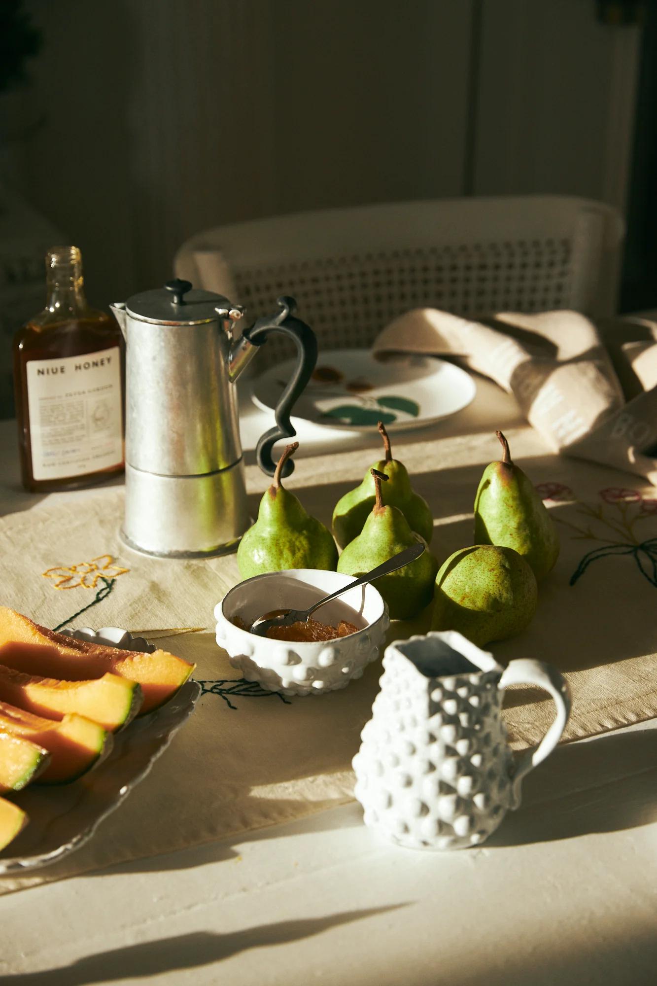 Cosy holiday breakfast spread on cream tablecloth. Pears, pitcher, sugar bowl, cantaloupe, coffee pot, honey, and rattan chair visible.