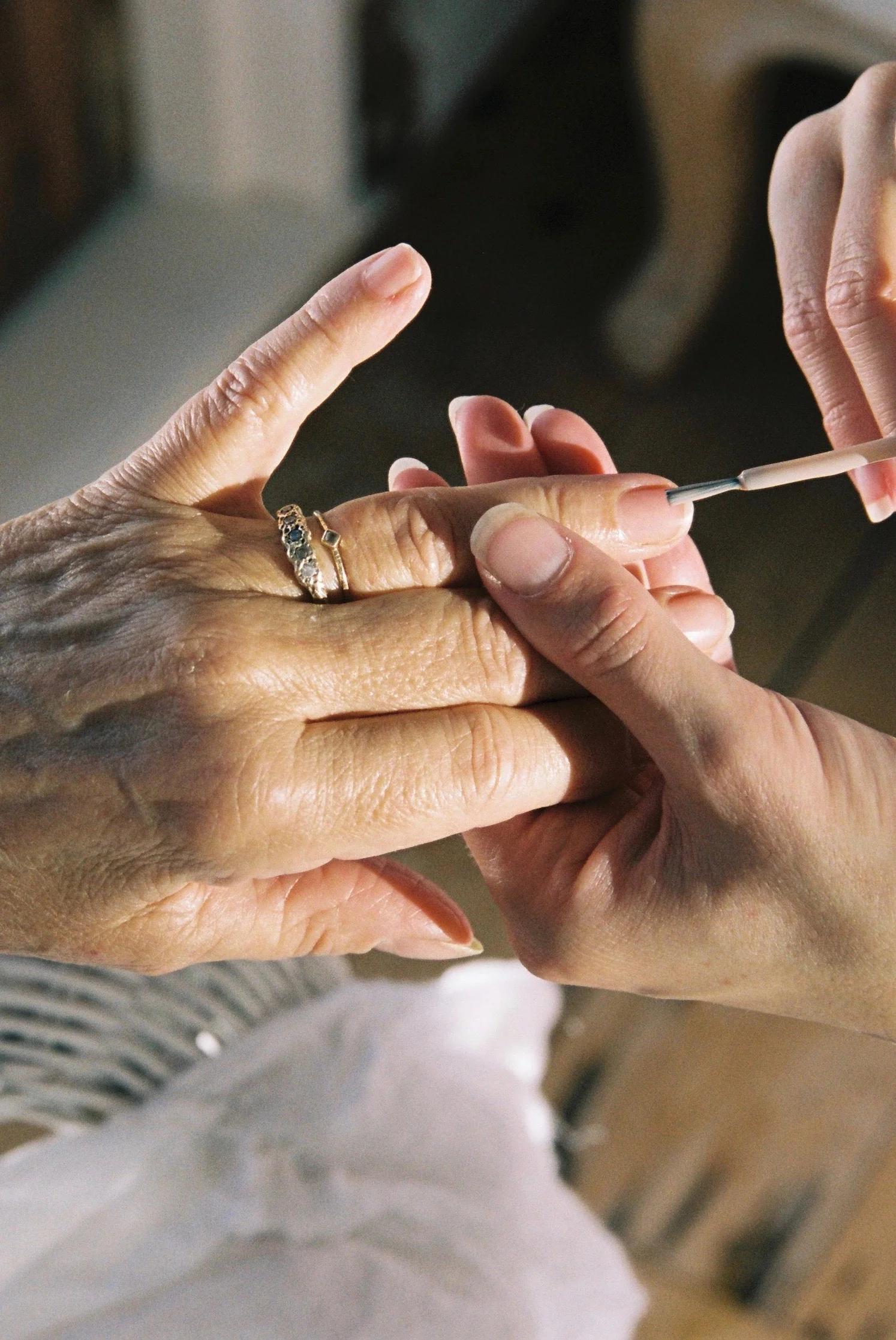 Close-up of two hands during a manicure. One older hand with a wedding ring is being carefully polished by a younger hand, indicating detailed and delicate nail care.