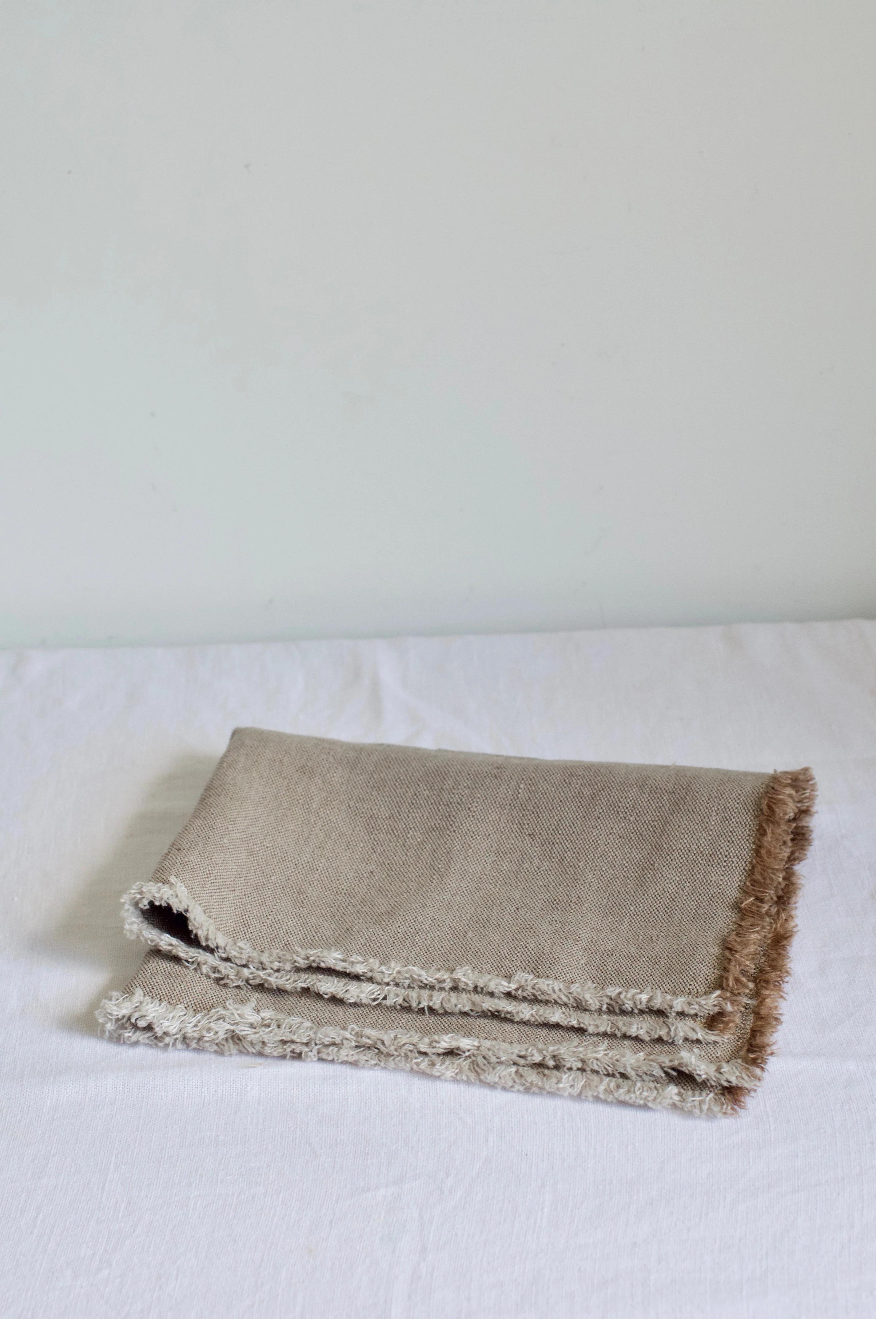 Folded beige blanket with fringed edges on white surface. Soft texture visible. Contemporary design touch.