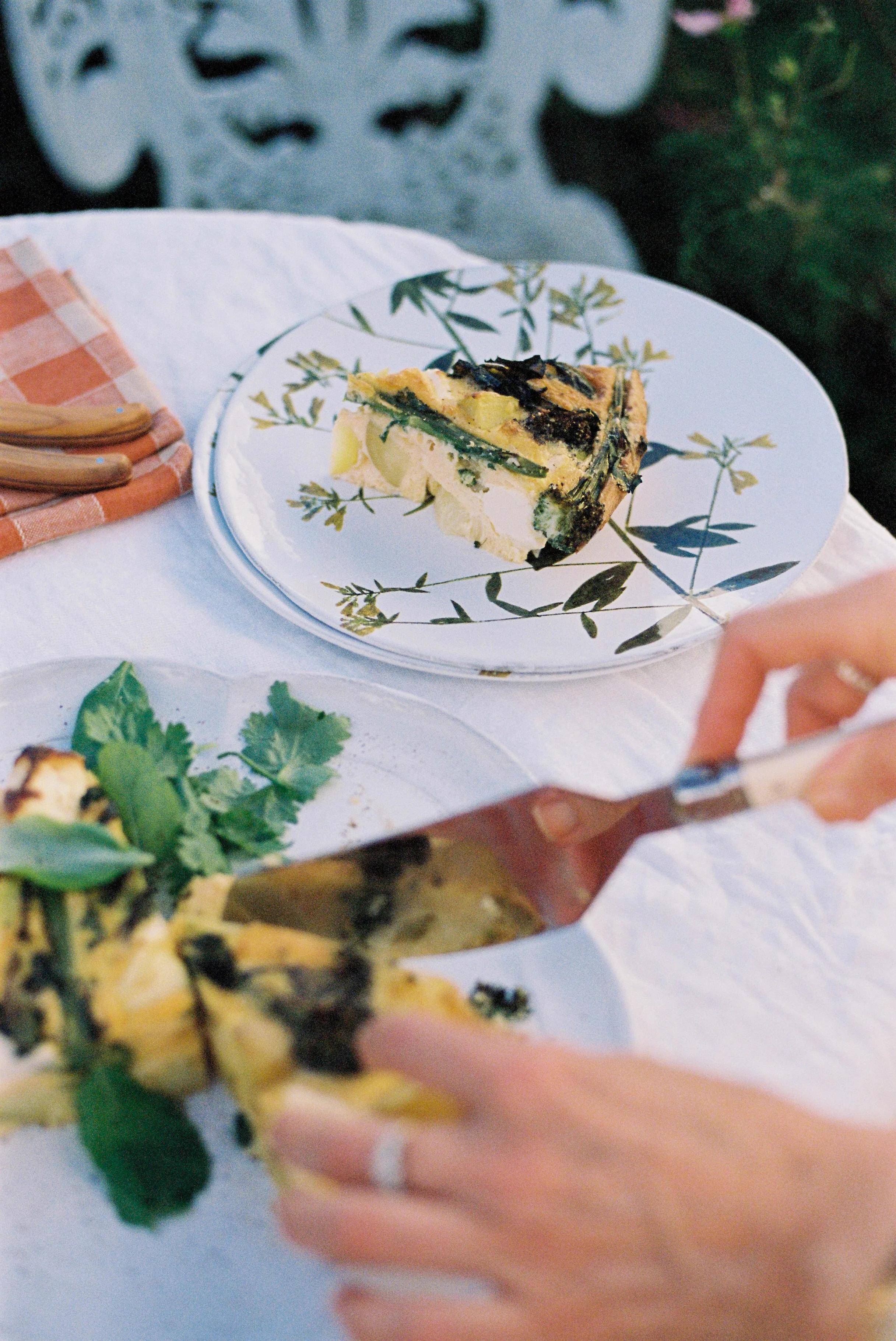 Person serving savoury tart in sunny garden. Slices on floral plates with orange-checked napkin. Tart garnished with herbs, decorative chair visible.