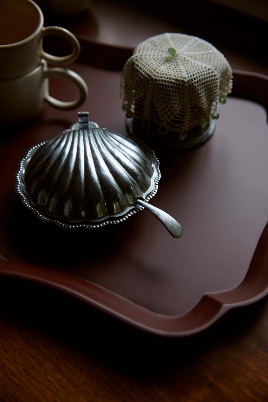 A silver dish with lid on a brown tray, alongside a jar covered in crochet and stacked cups. The warm lighting creates a cosy, vintage atmosphere.