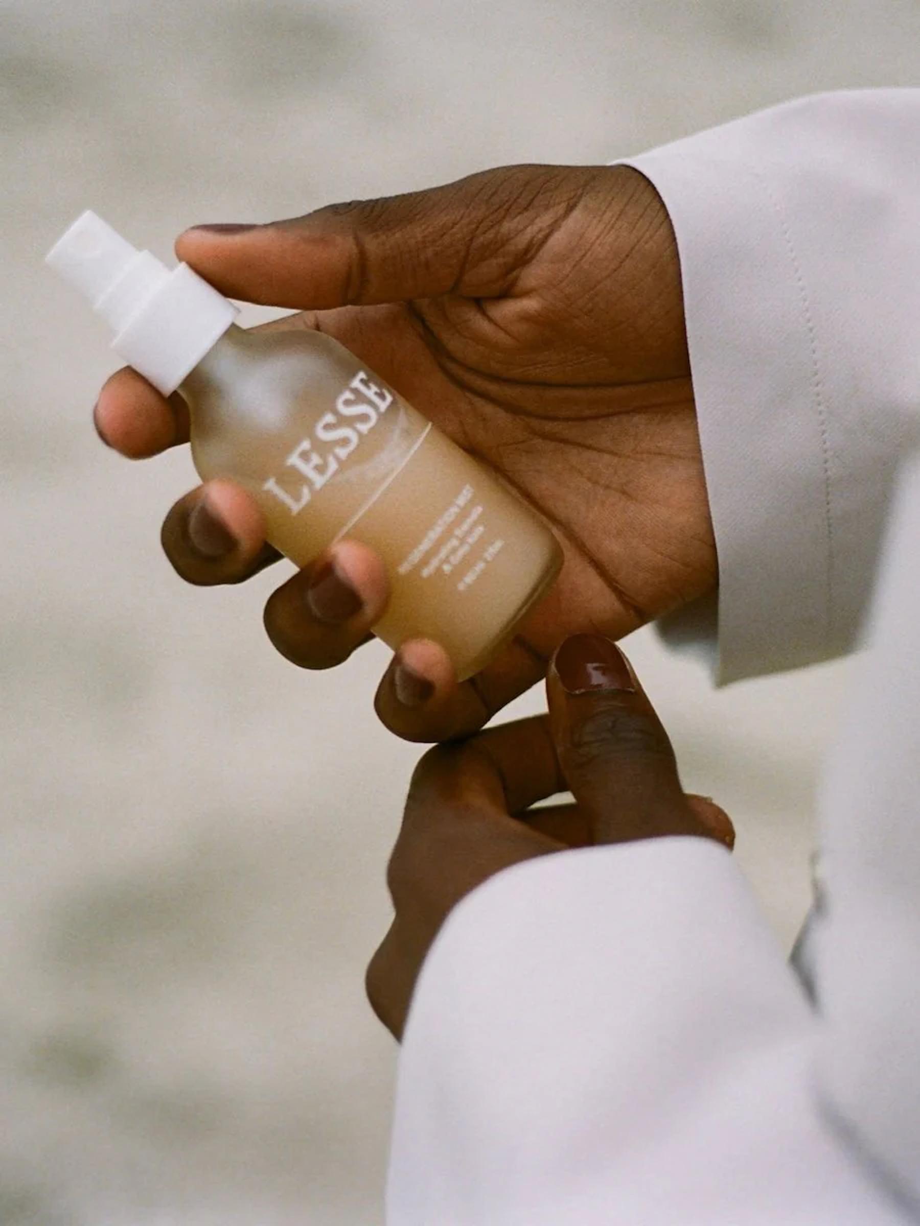 Dark-skinned hands hold LESSE spray bottle. White-labelled bottle contrasts with light sleeve. Blurred background.