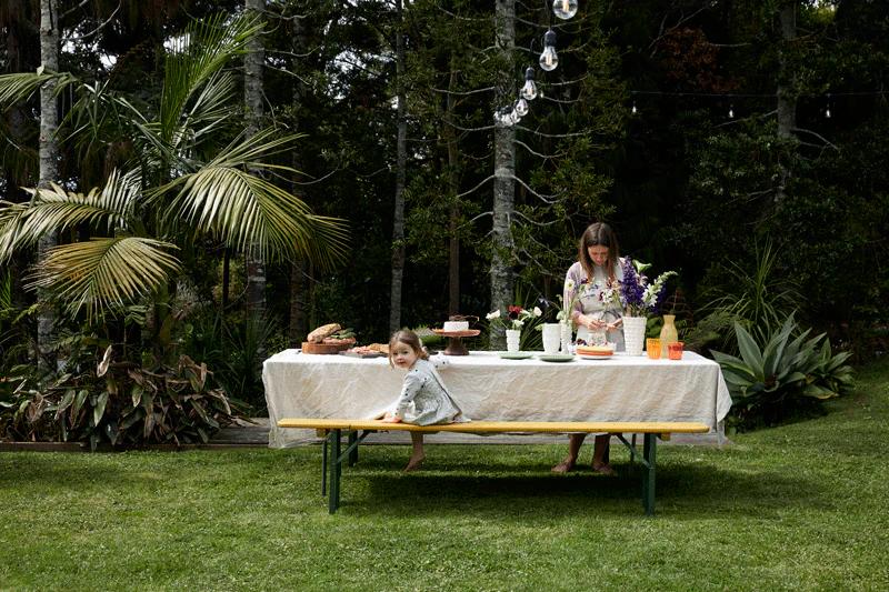 Rachel Carley and a child are at an outdoor table set for a dinner party. The child sits on a yellow bench, while Rachel arranges food and flowers, surrounded by greenery and hanging lights.