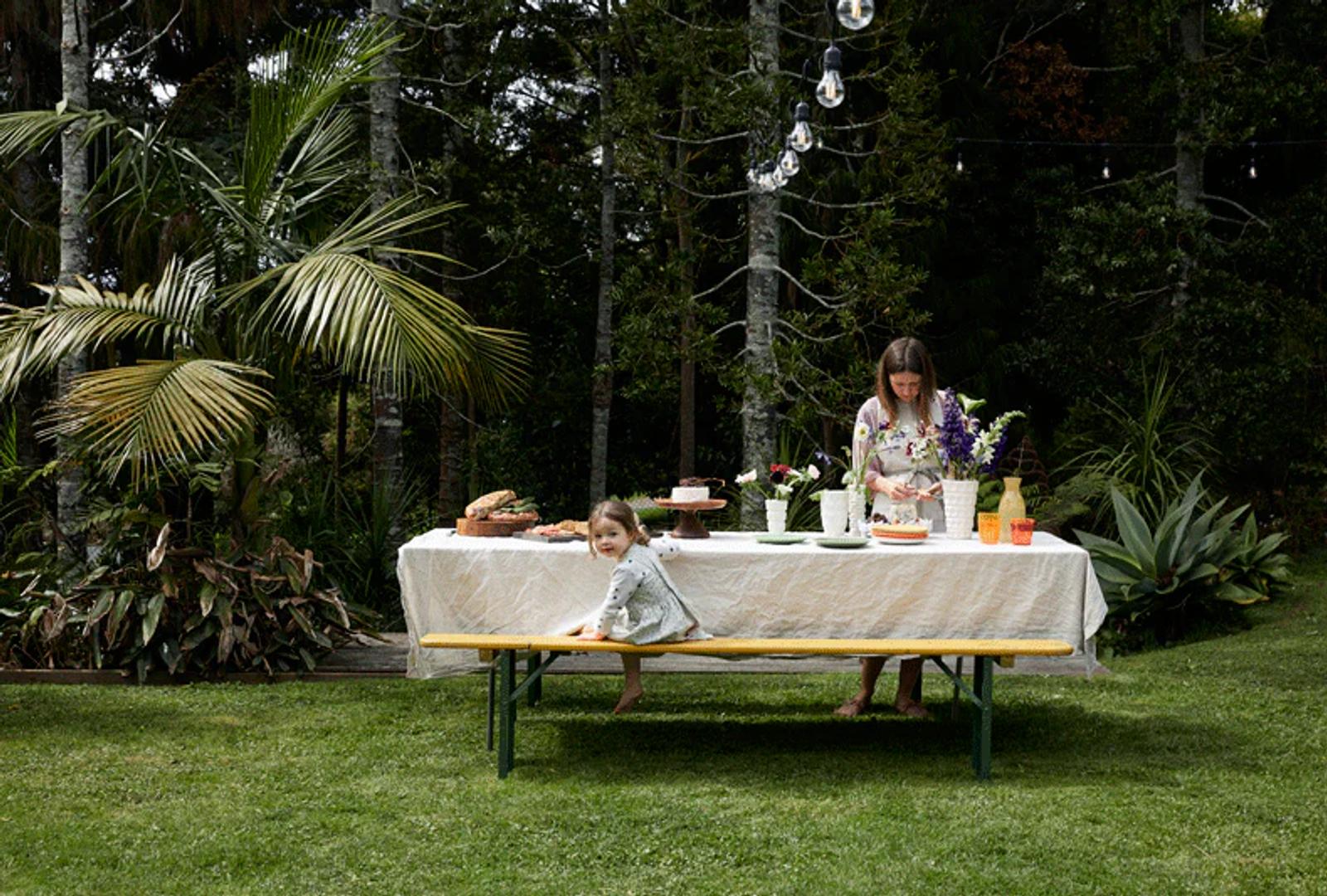 Rachel Carley and a child are at an outdoor table set for a dinner party. The child sits on a yellow bench, while Rachel arranges food and flowers, surrounded by greenery and hanging lights.
