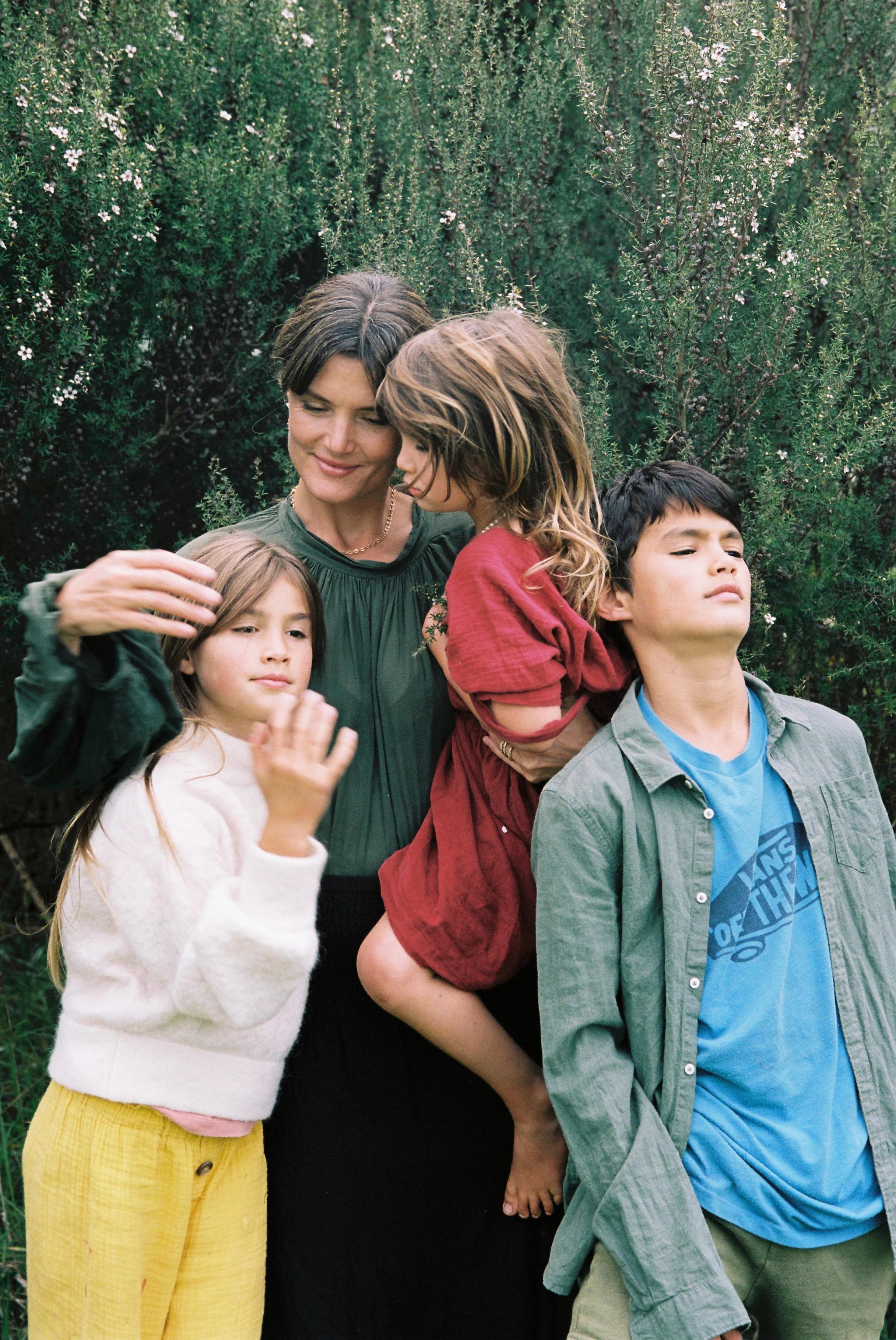 A woman stands with three children in front of green foliage. She hugs a girl in a white sweater and yellow pants, holds a child in a red dress, and stands beside a boy in a blue shirt.