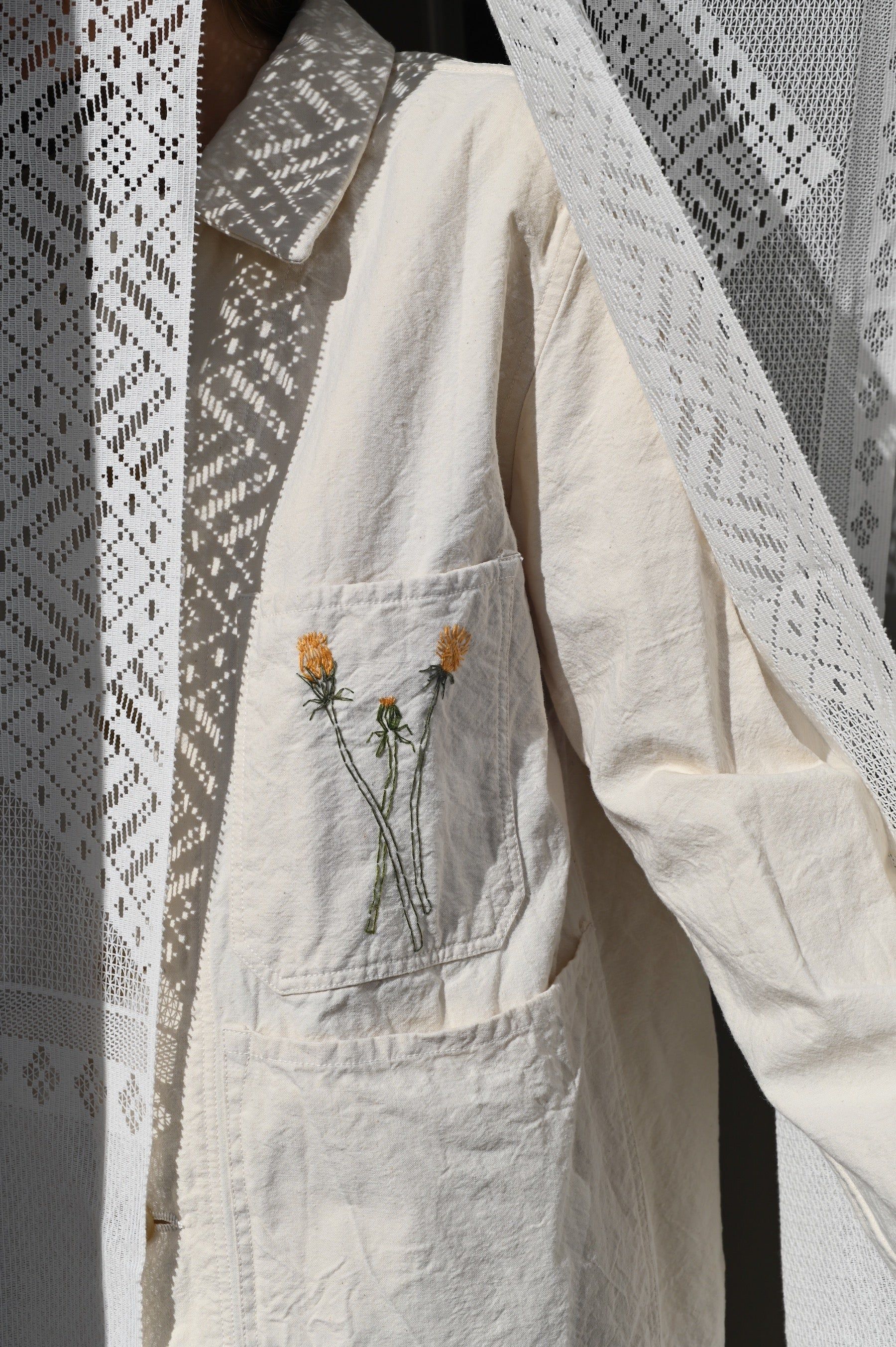 Cream shirt with embroidered dandelions by Singular Garments. Lace curtain casts geometric shadows on fabric.