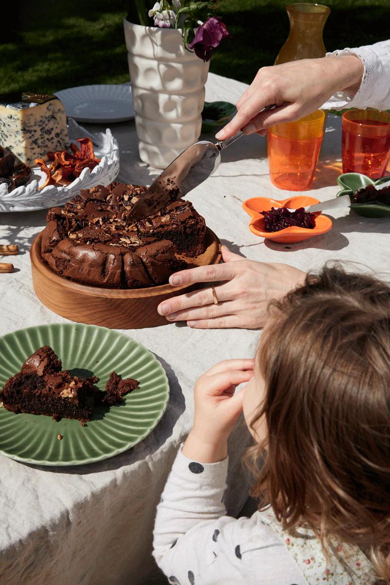 Person slices nut-topped cake as eager child watches. Table set with cheese, dried tomatoes, and orange glasses under sunlight.