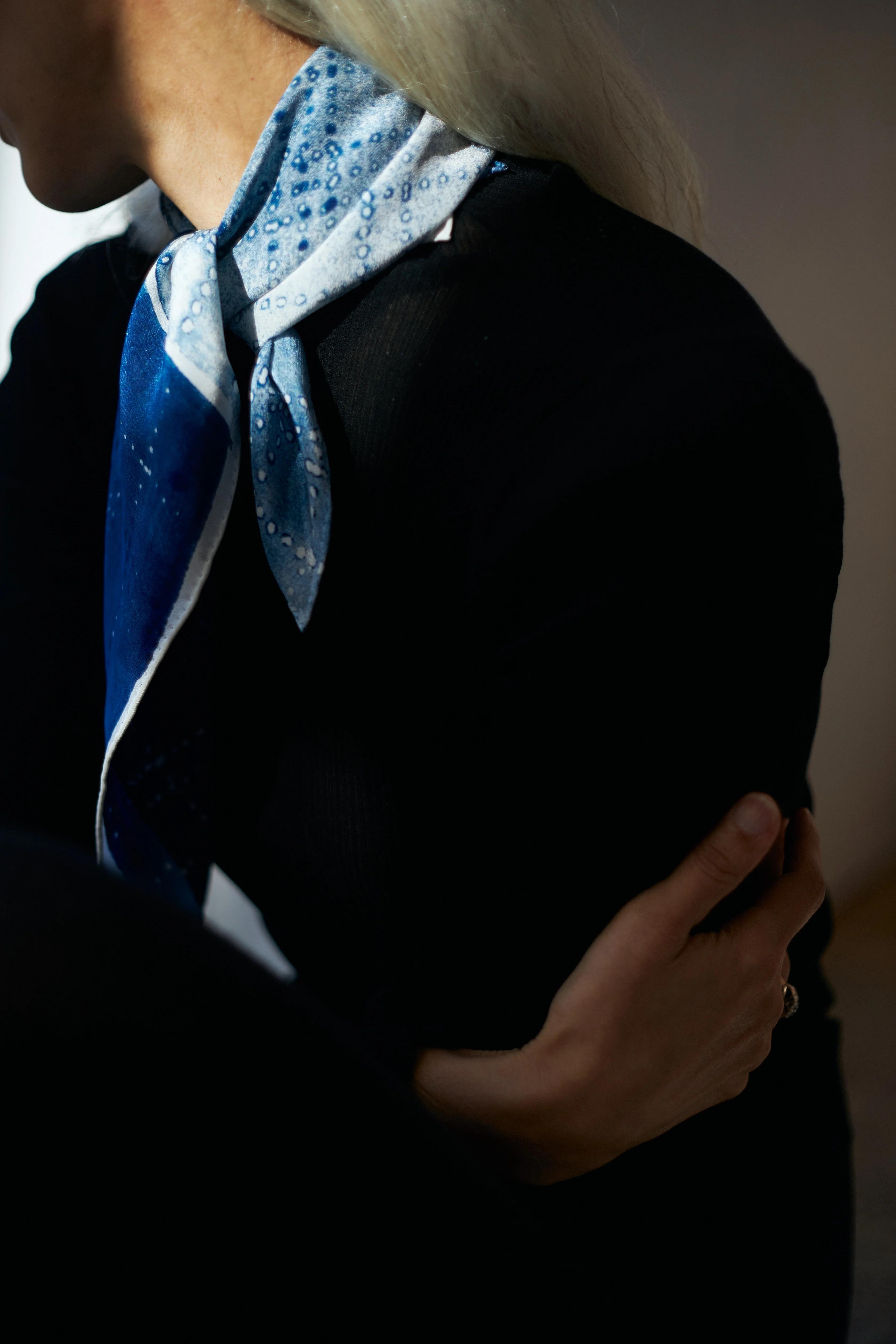 A person wearing a dark long-sleeve top and a blue patterned scarf around their neck, reminiscent of contemporary art textile design, is sitting with their left arm crossed, holding their right arm. The background is dimly lit, creating an intimate ambiance.