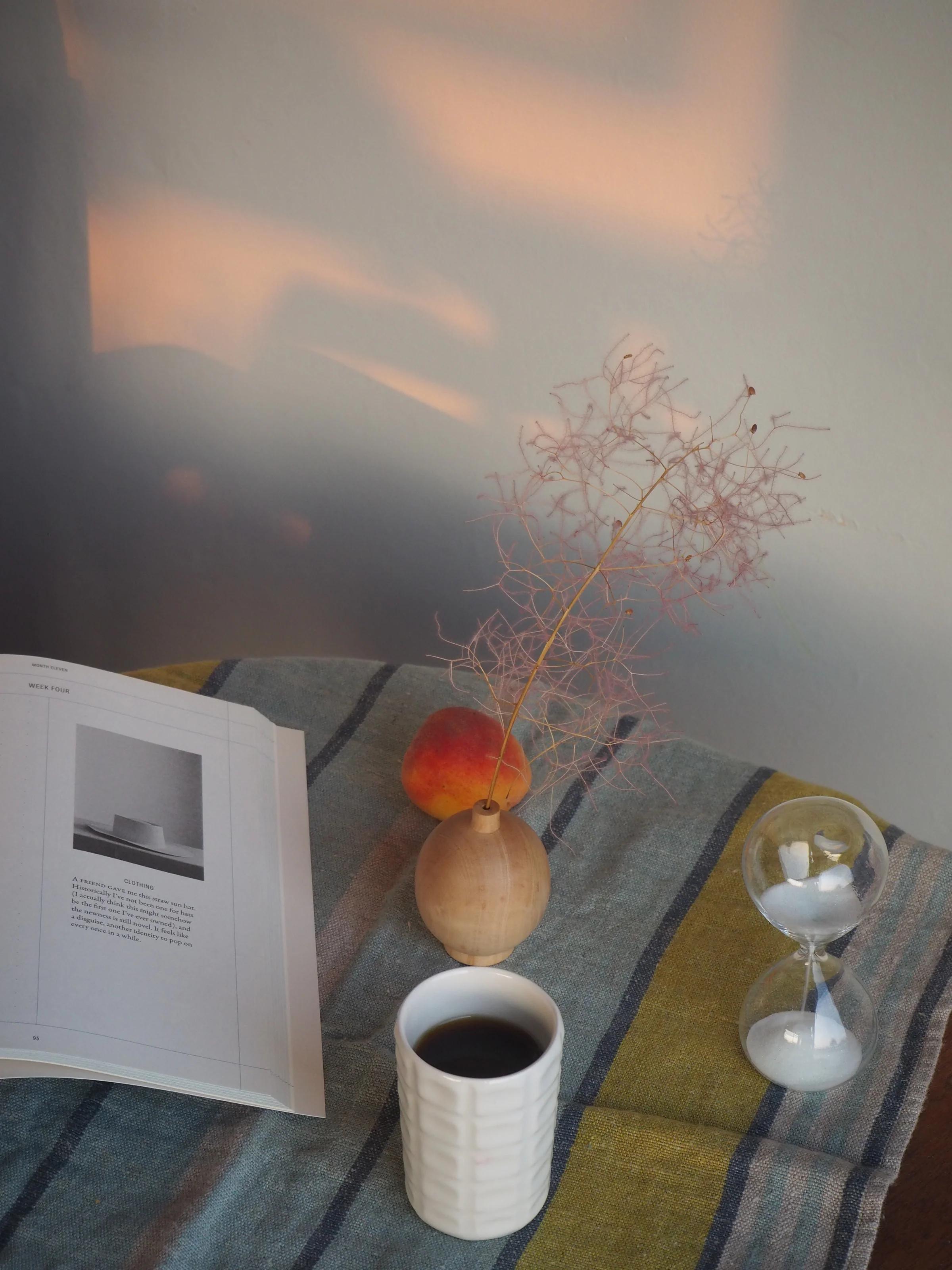 A table with an open book, apple, branch, coffee cup, and hourglass on a striped cloth, illuminated by soft sunlight for a cozy atmosphere.