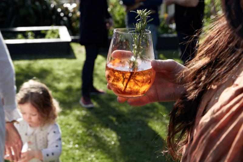 Person with rosemary-garnished red drink at outdoor party. Child playing in background. Sunlit grassy area visible.